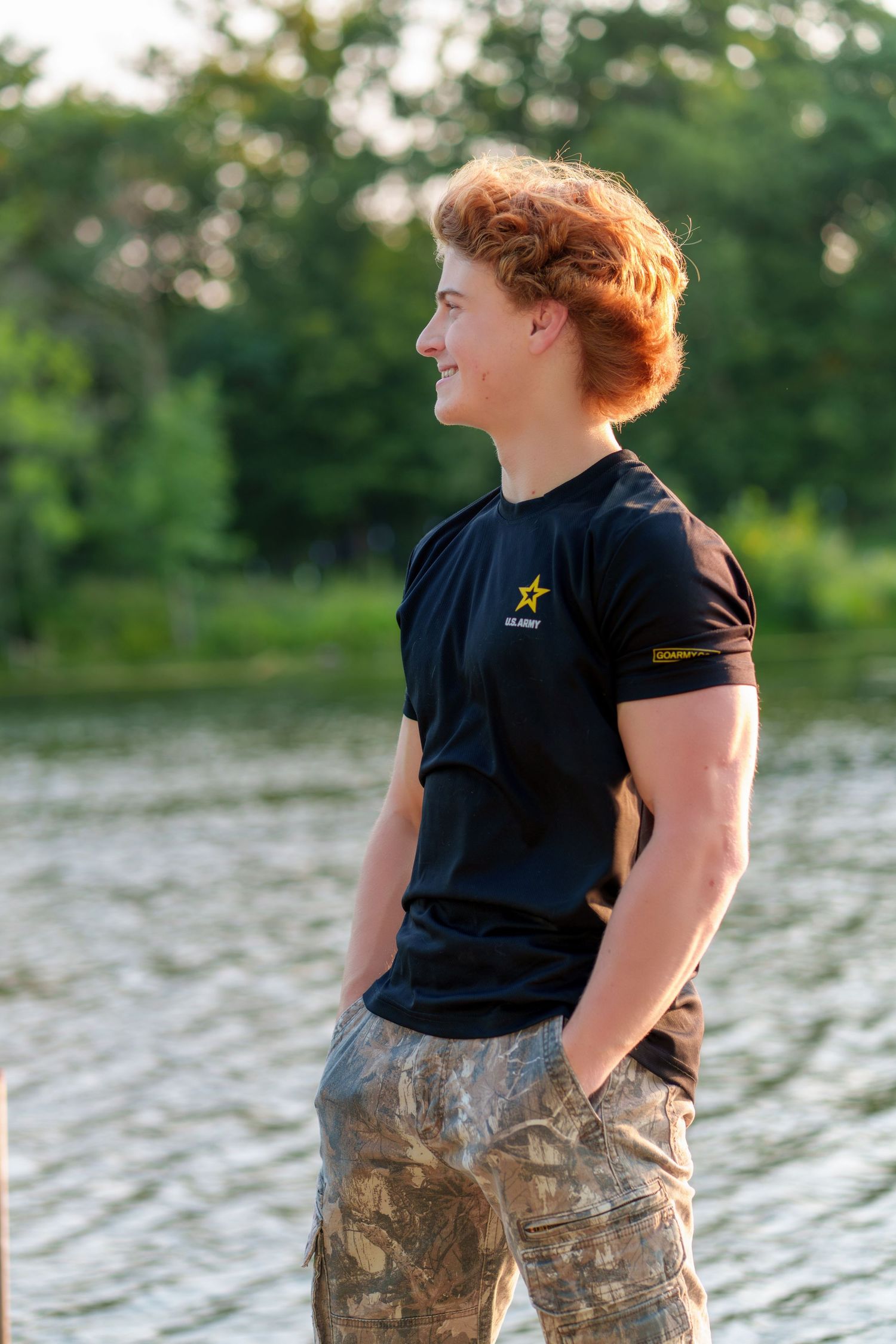 Young man with red hair wearing a black Army t-shirt and camo shorts standing near a lake outdoors.