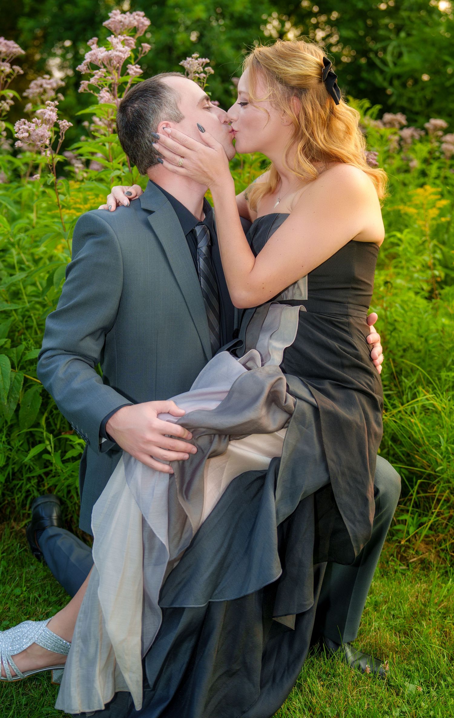 Couple sharing a romantic kiss outdoors, man in gray suit holding woman in elegant black gown among lush green foliage.