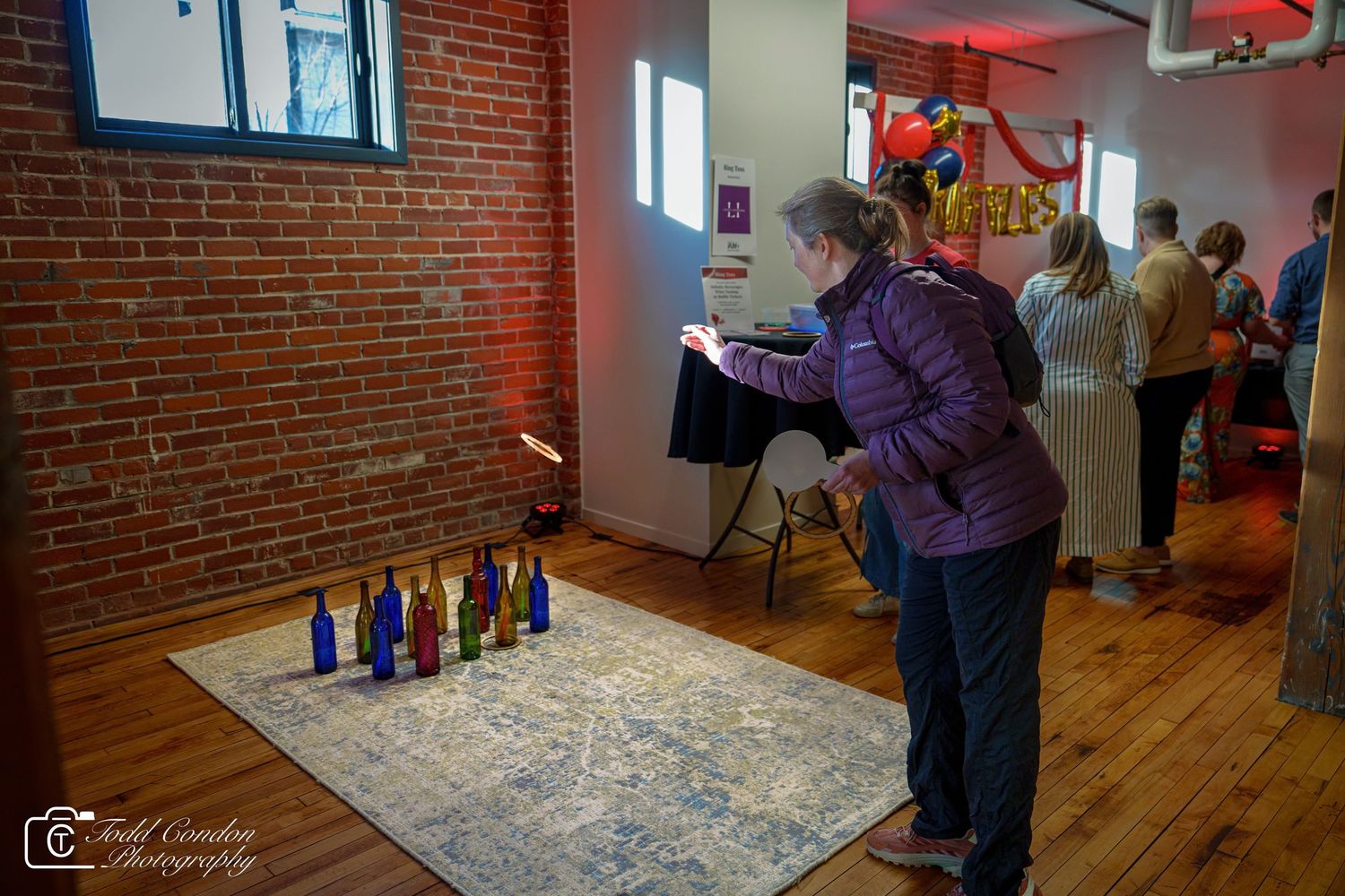 Woman lighting colorful glass bottles on a rug at an indoor event in a brick-walled venue.