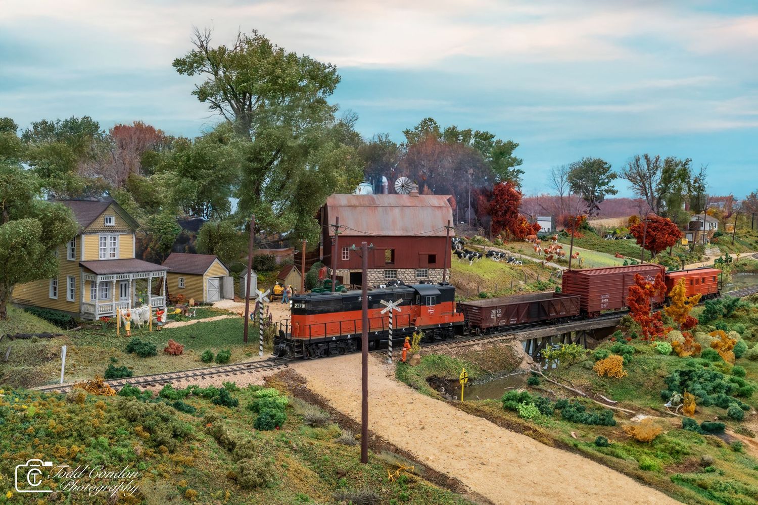 Model railroad layout featuring autumn scenery with trees, buildings, and orange locomotives on a detailed track system.