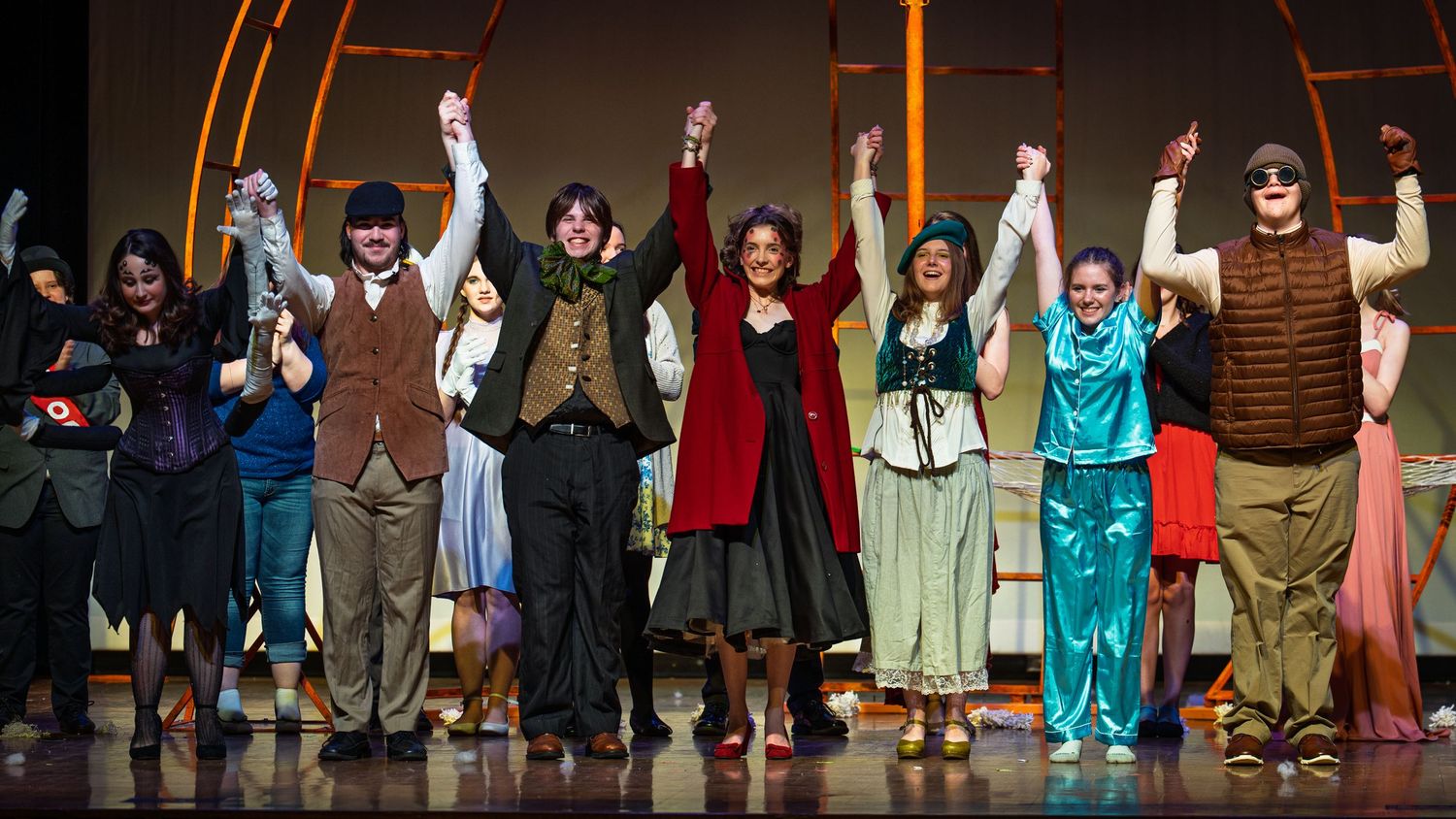 Theater performers take a bow at curtain call on stage with raised arms in colorful costumes.