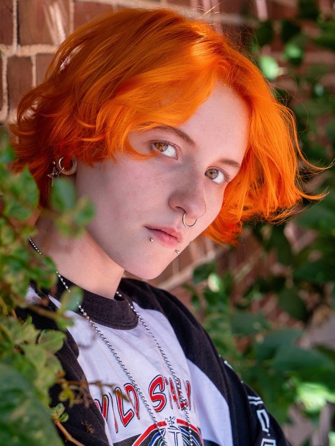 A person with vibrant orange hair wearing a black and white shirt poses against a leafy green background.