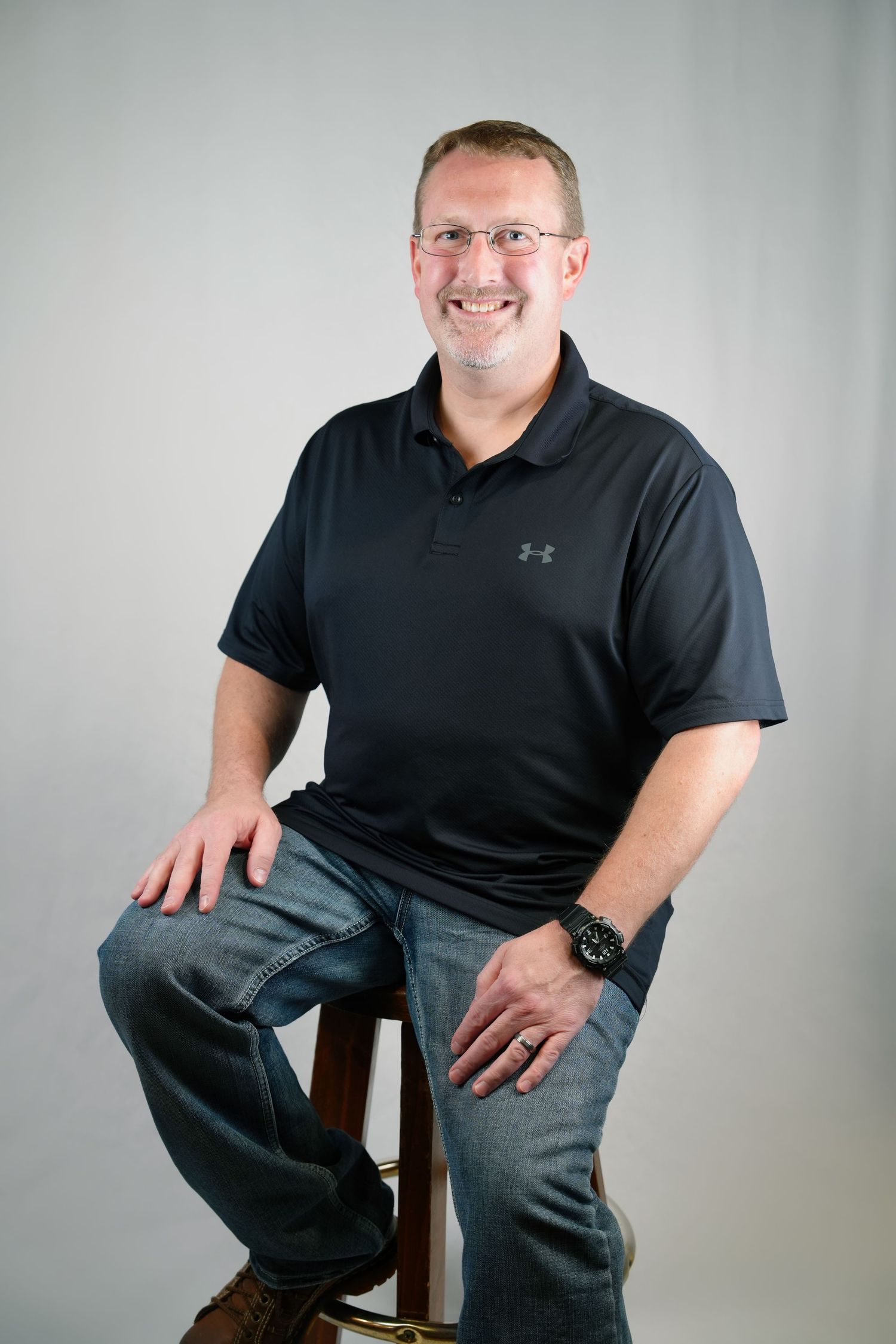 Professional studio portrait series showing person in black Under Armour polo shirt seated on wooden stool.
