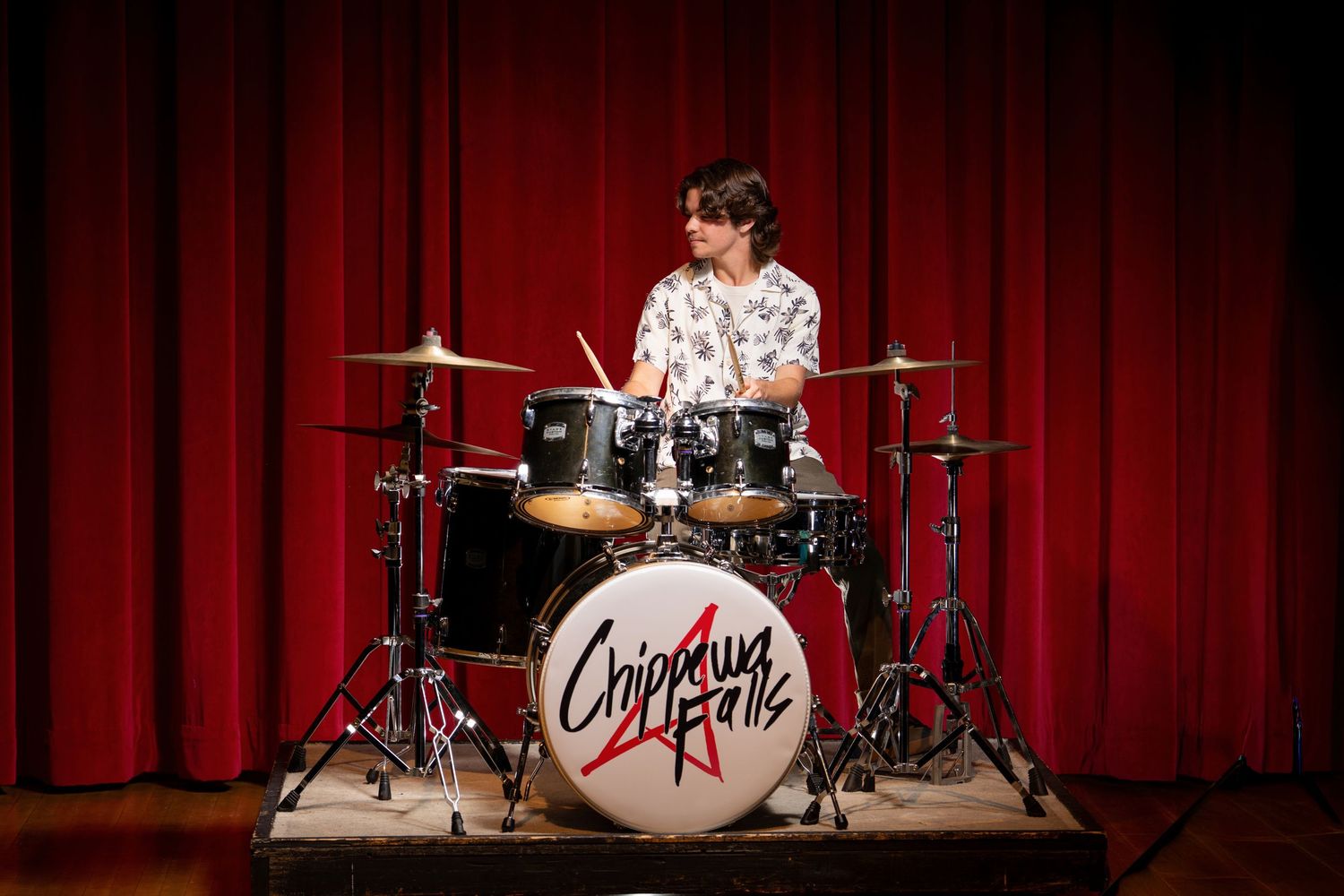 Black drum kit with red curtain backdrop on stage illuminated by dramatic lighting.
