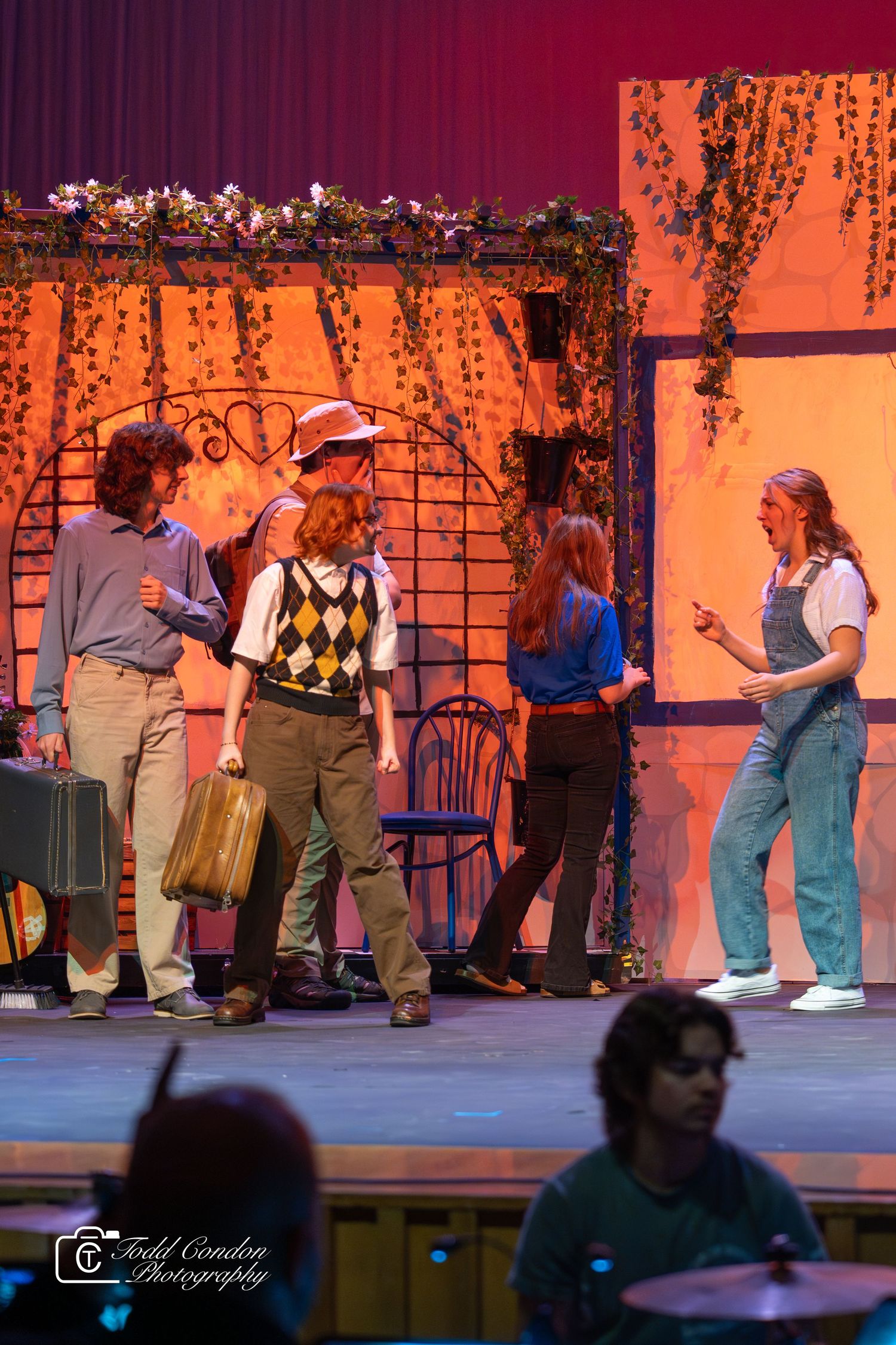 Performers on stage in a theatrical production with orange lighting and dark silhouettes moving across the scene.