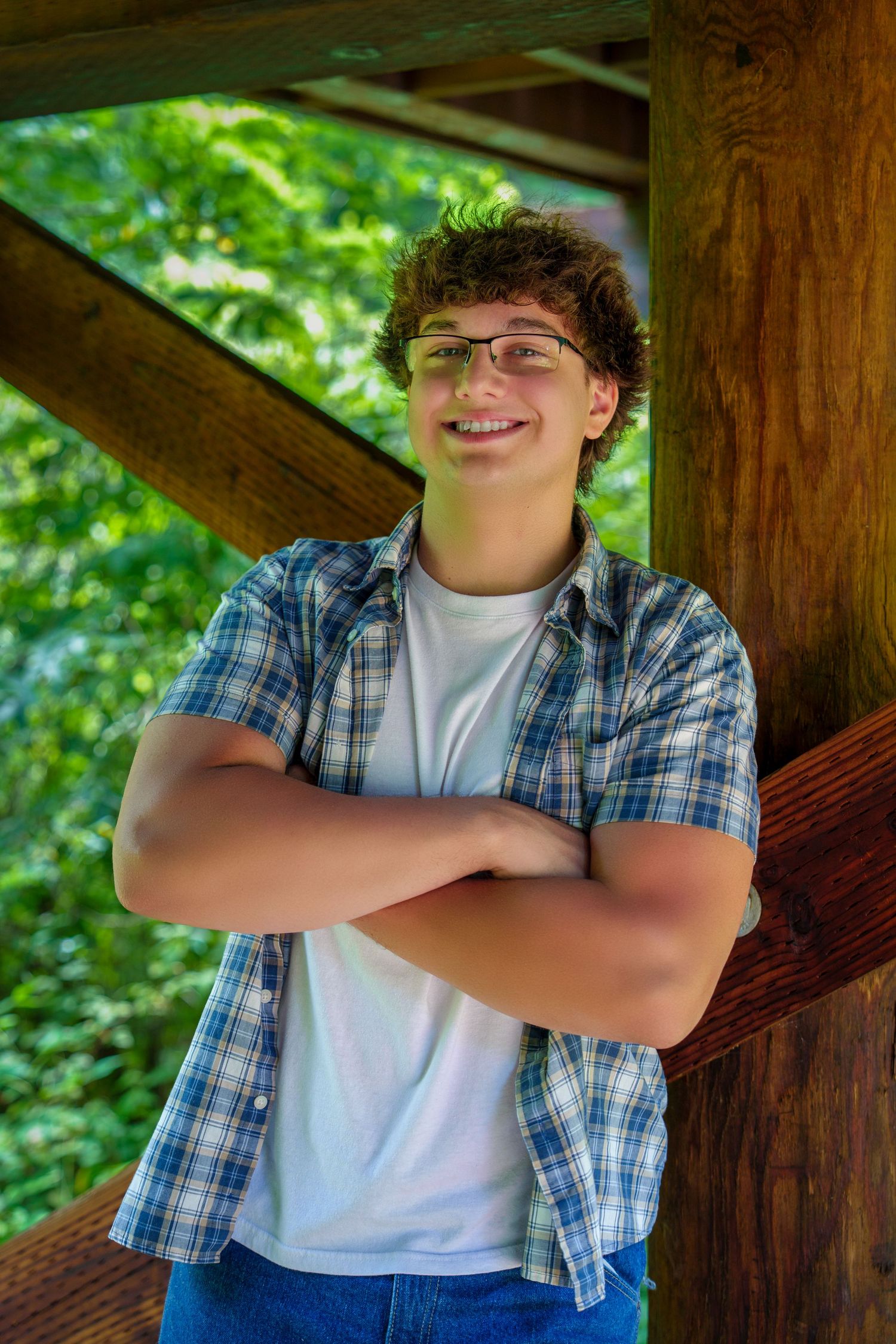 Smiling teenage boy with curly hair and glasses, arms crossed, wearing plaid shirt, leaning against wooden structure outdoors.
