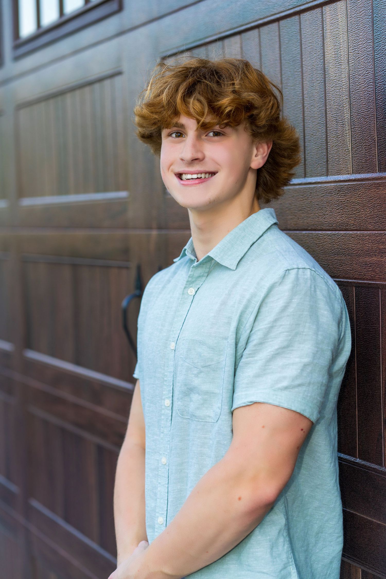 Smiling teenage boy with curly auburn hair wearing a light blue shirt, leaning against a wooden garage door.