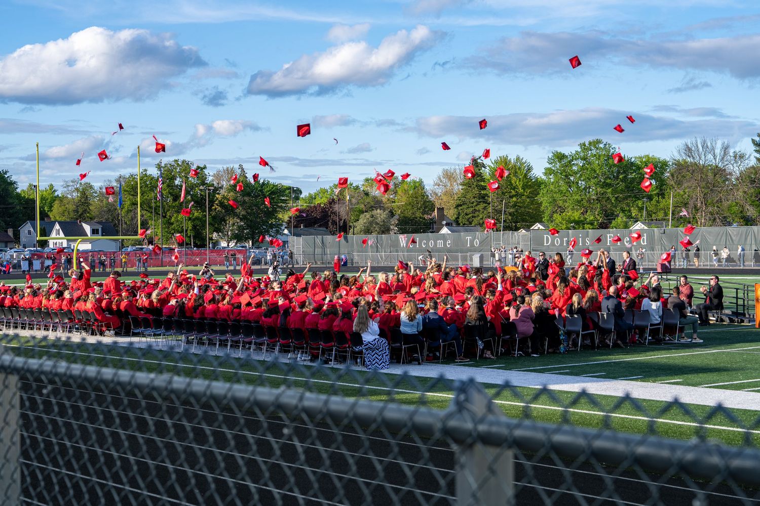 Graduates in red caps and gowns toss mortarboards into the air during an outdoor graduation ceremony on a school athletic field.