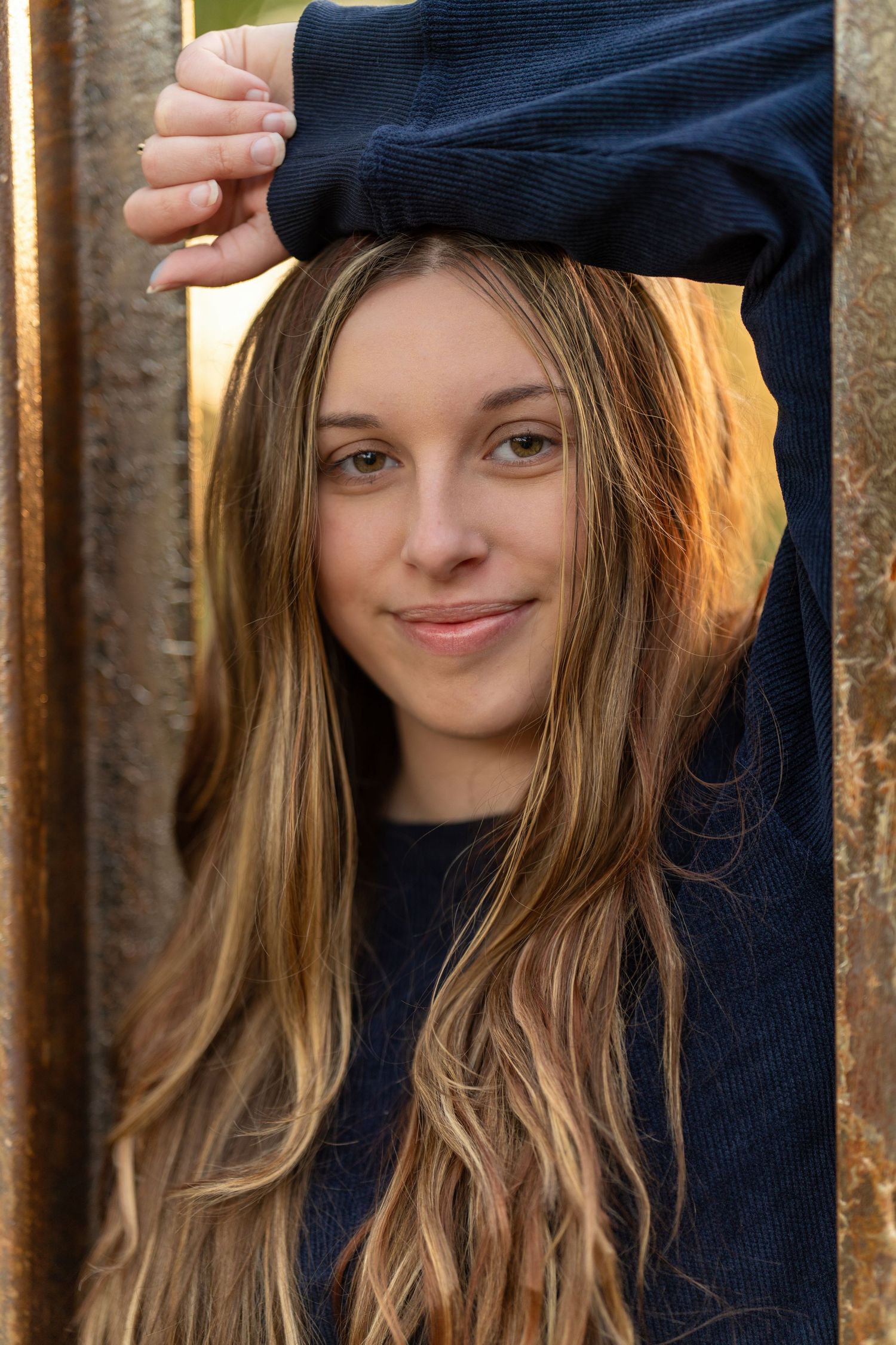 A person wearing a navy blue sweater smiles warmly while posing against a rustic background.