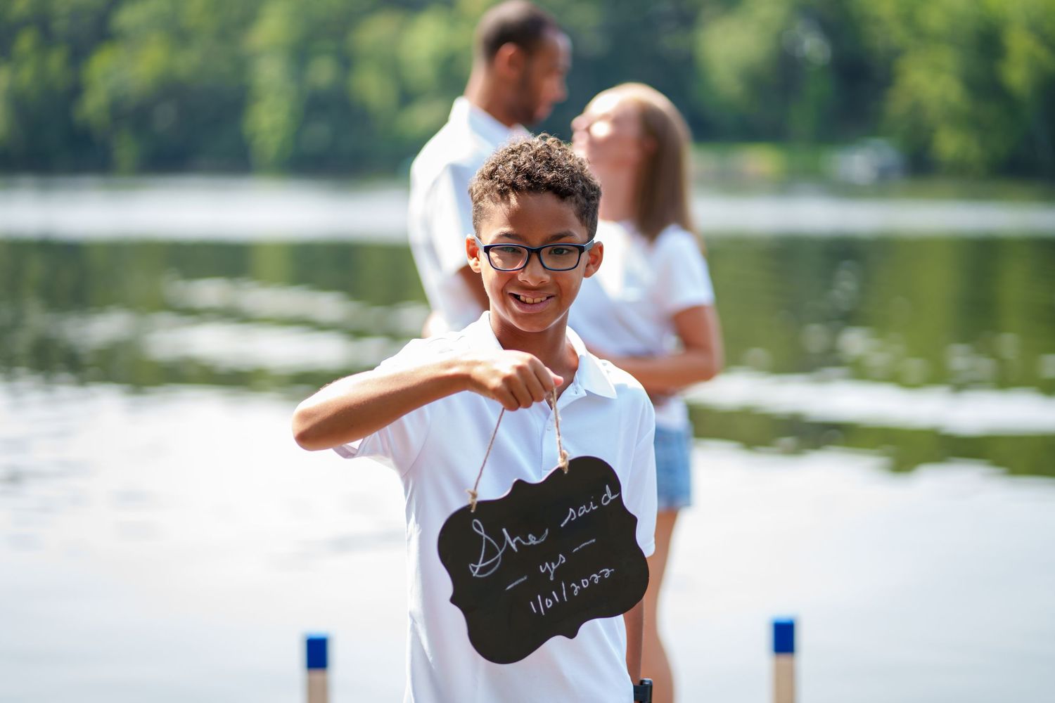 Young boy in white polo shirt smiles and poses near a lake during a sunny summer day.