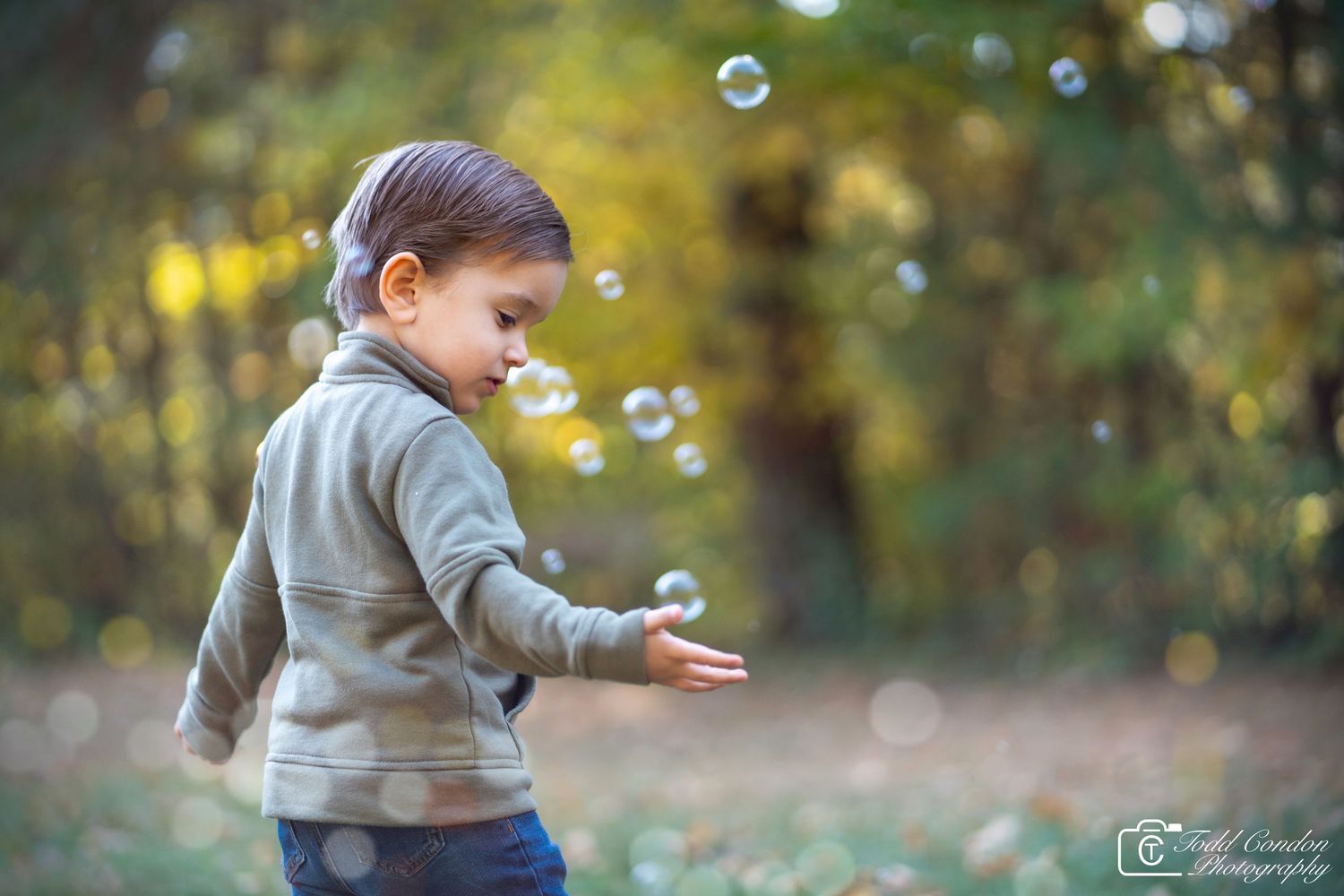 A child in a gray sweater plays with floating bubbles in a fall outdoor setting.
