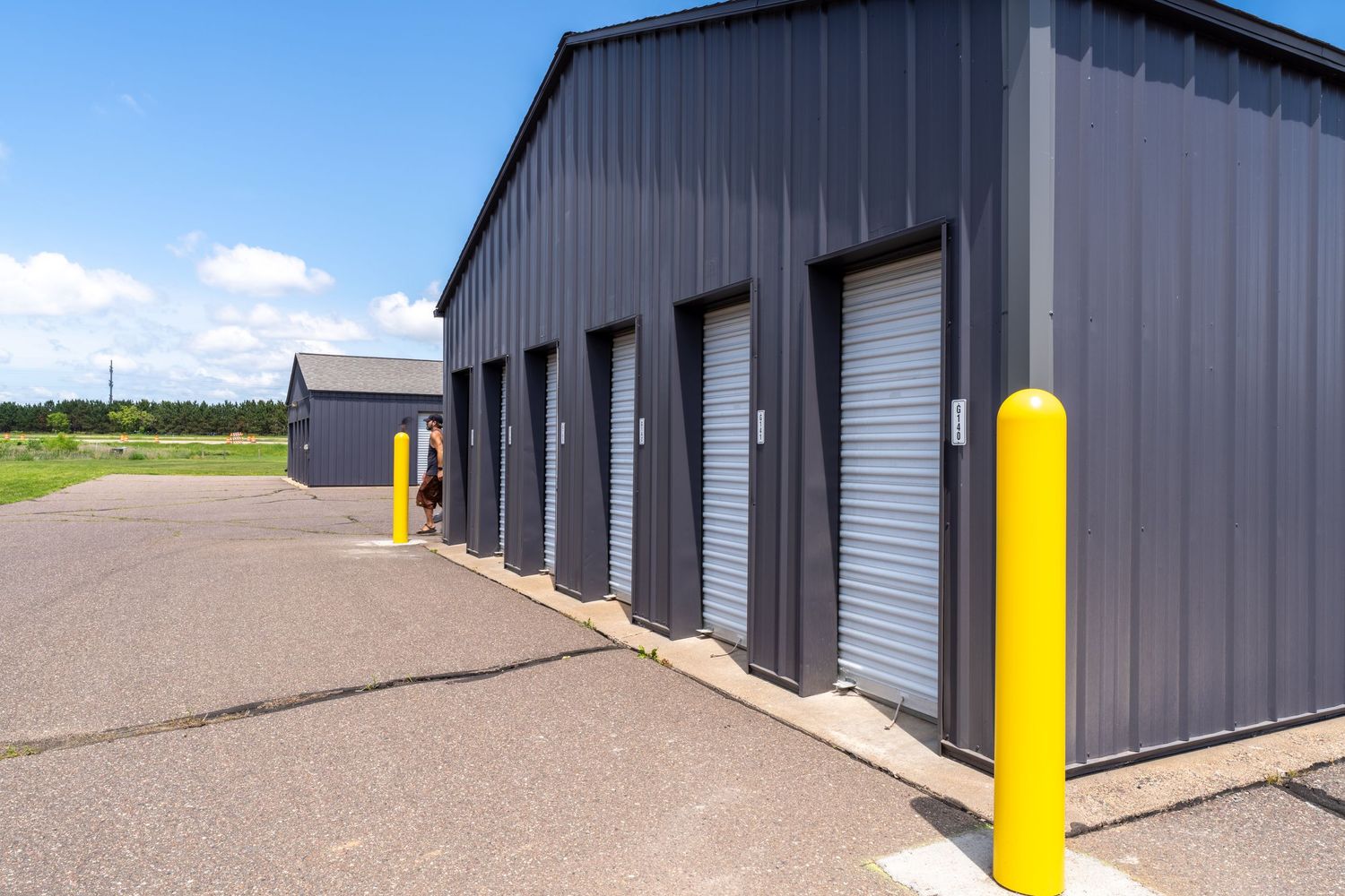 A row of dark gray self storage units with bright yellow safety bollards along a concrete driveway.