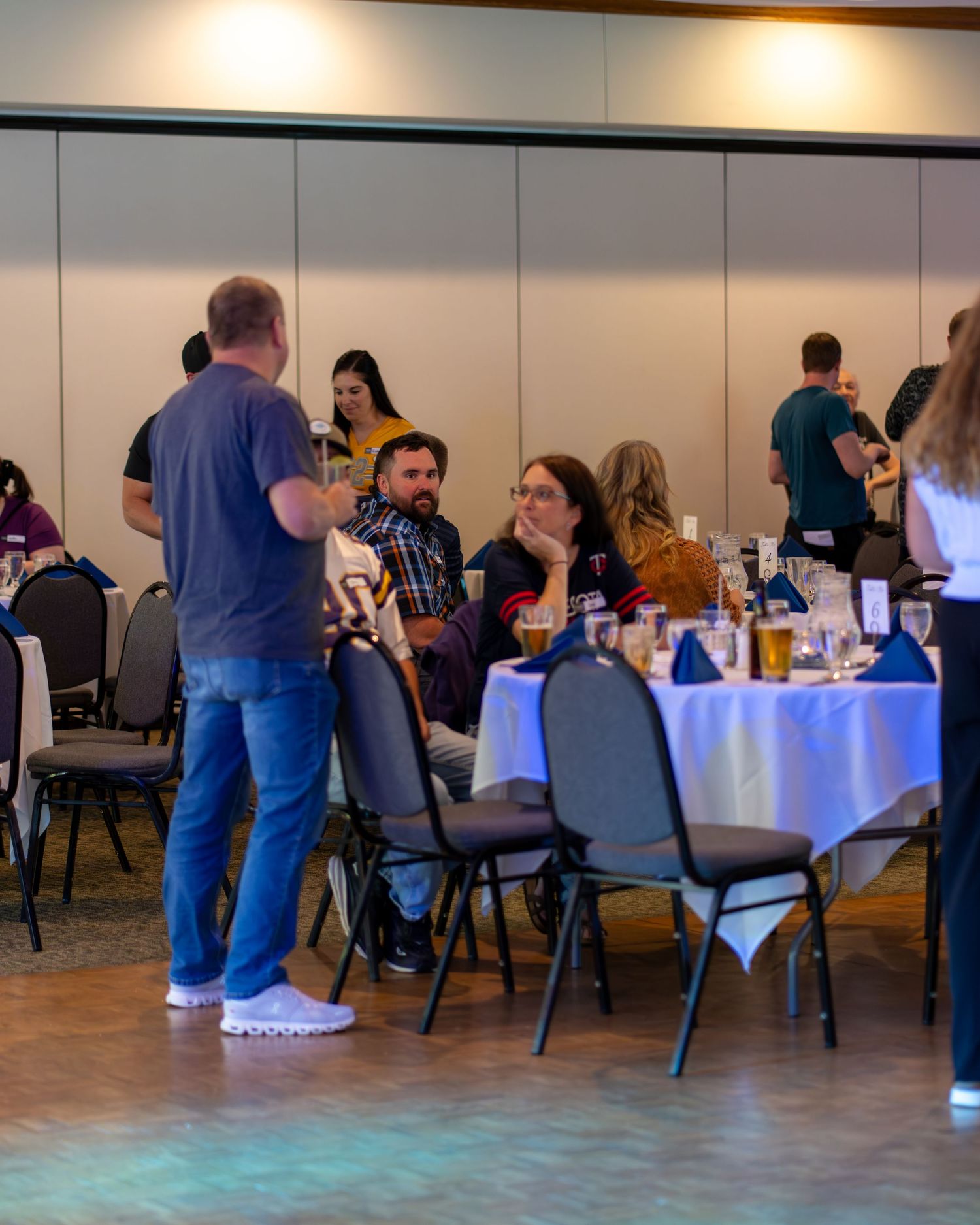 People gather at round tables with white tablecloths in a banquet hall setting for a social event.