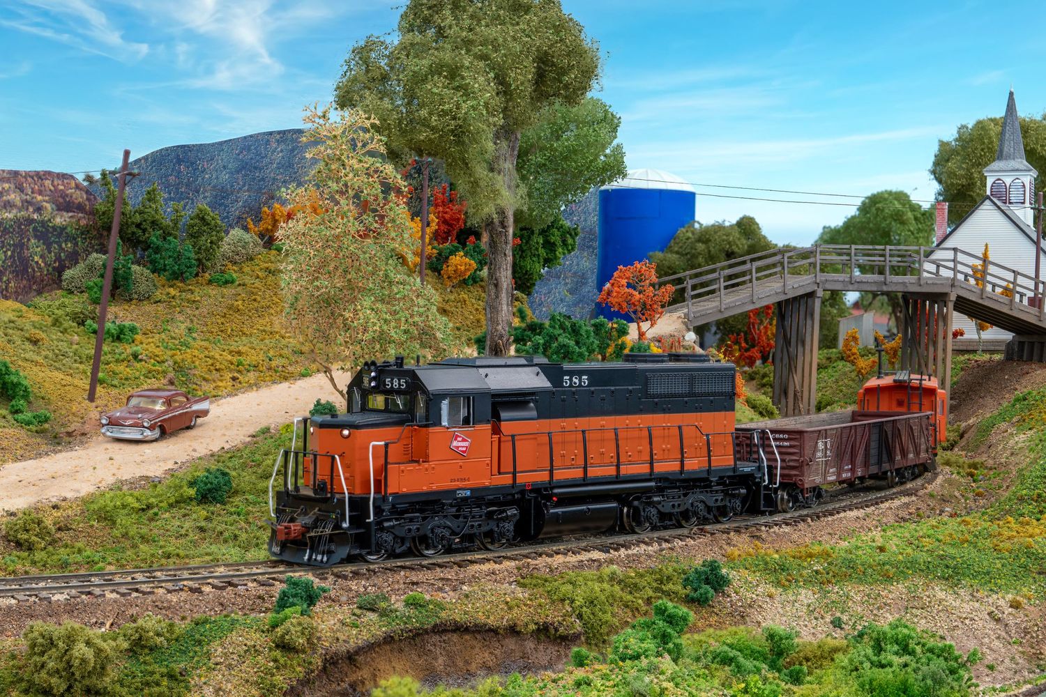 A vintage freight train with orange cargo cars winds through lush green countryside on a sunny day.