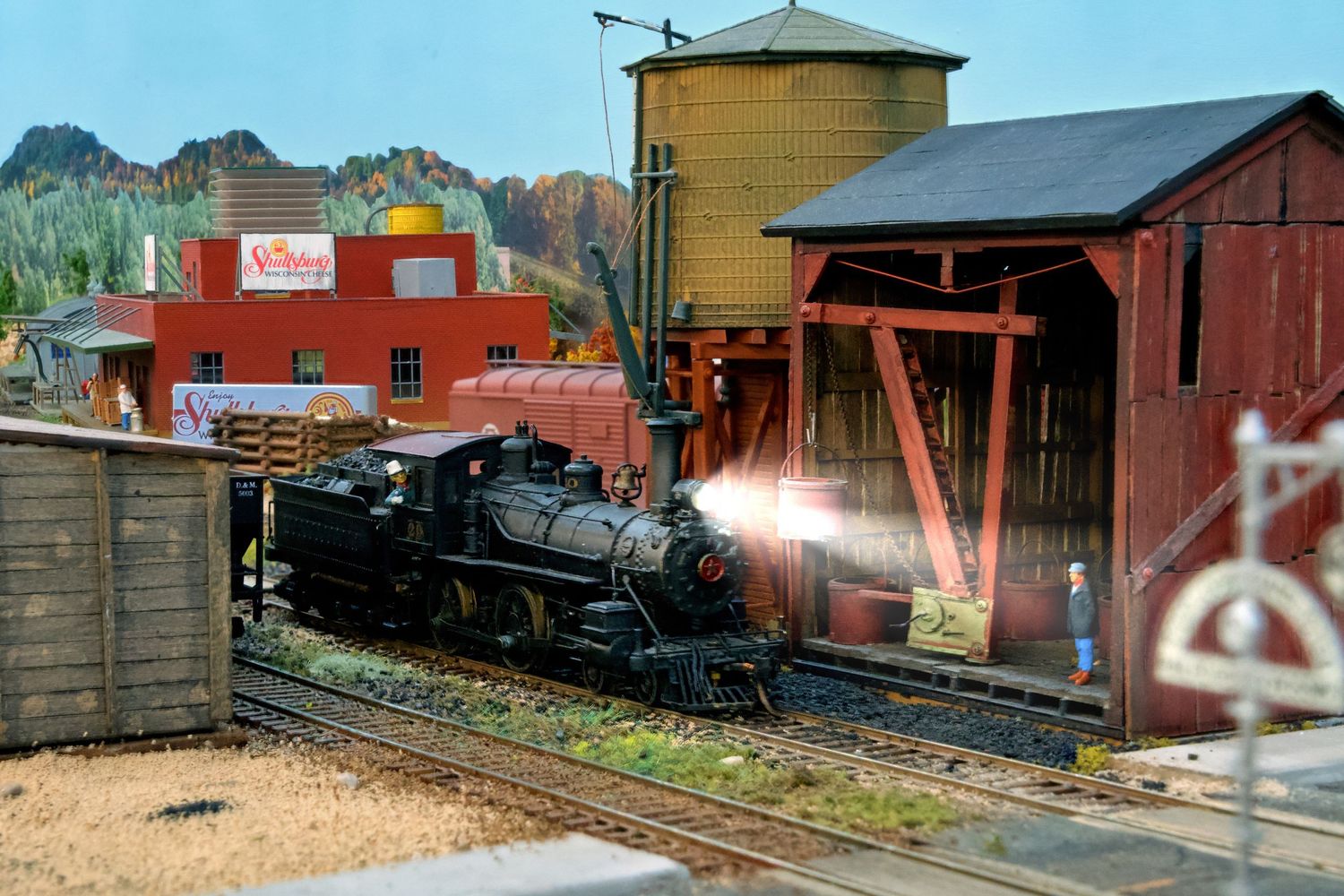 Vintage train cars and buildings line up along railroad tracks at a historic railway station yard.