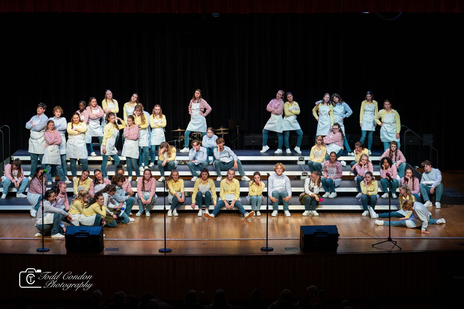 A large group of performers in light-colored outfits arranged on risers during a stage production.