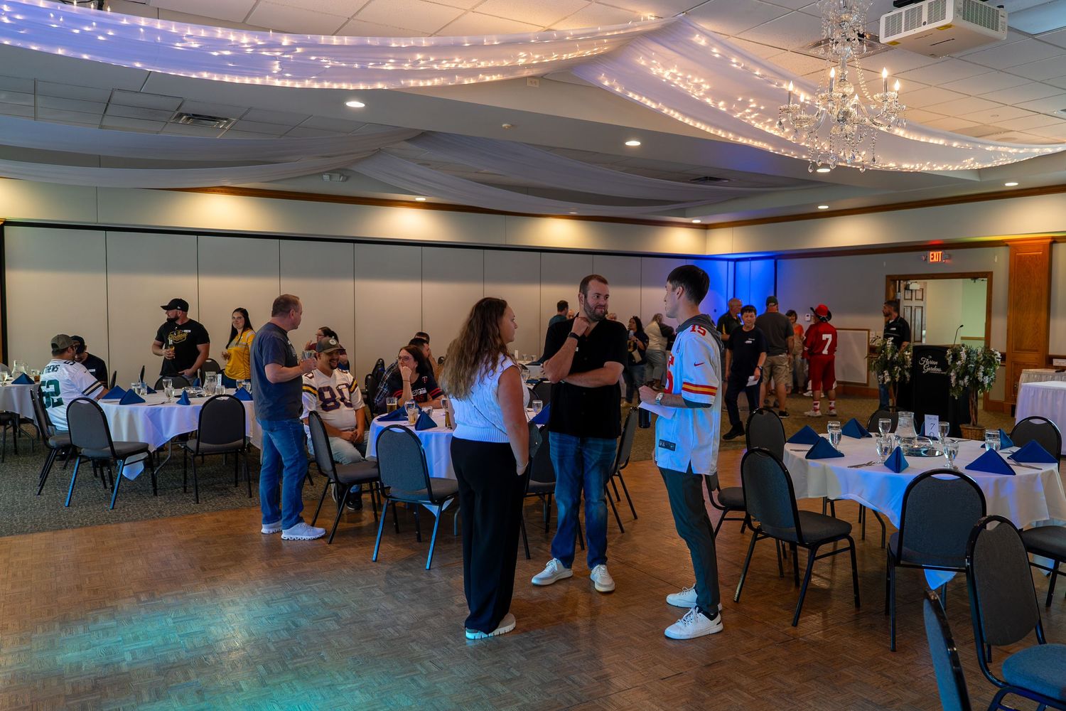 People gather in a large banquet hall with sparkling chandeliers and dark carpeting for an indoor event.