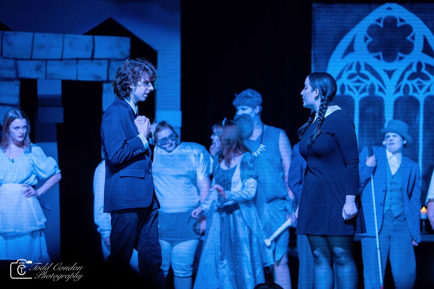 Theater performers in period costumes act in a dramatic scene bathed in blue stage lighting.