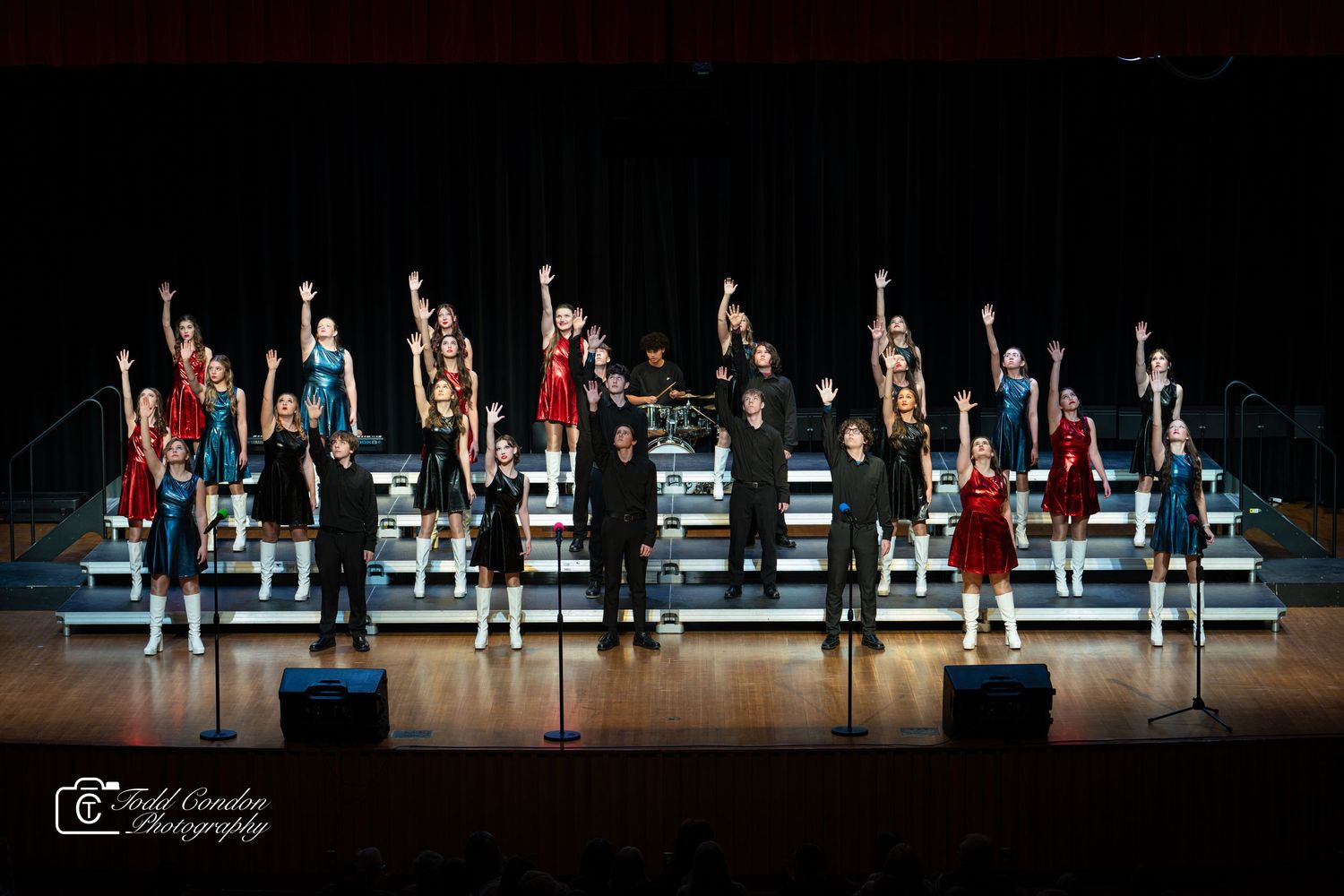 Show choir performers stand on risers in coordinated red and blue outfits during a stage performance.