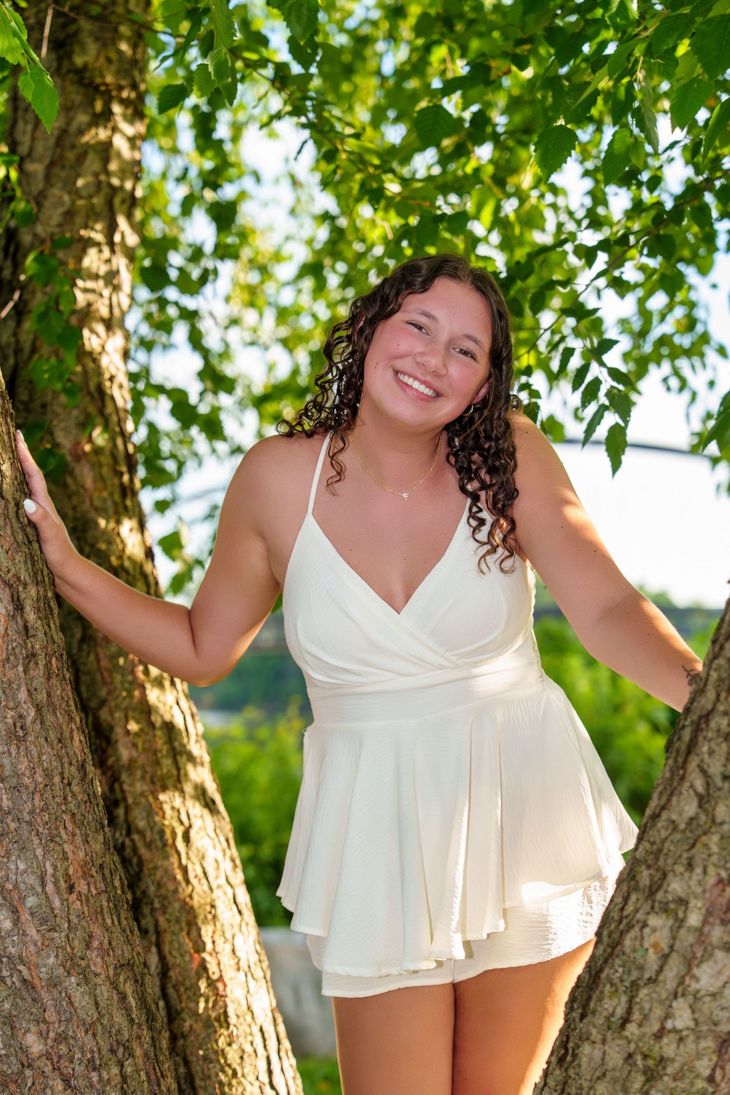 Young woman in white romper smiling between two trees with green leaves in a sunny outdoor setting.