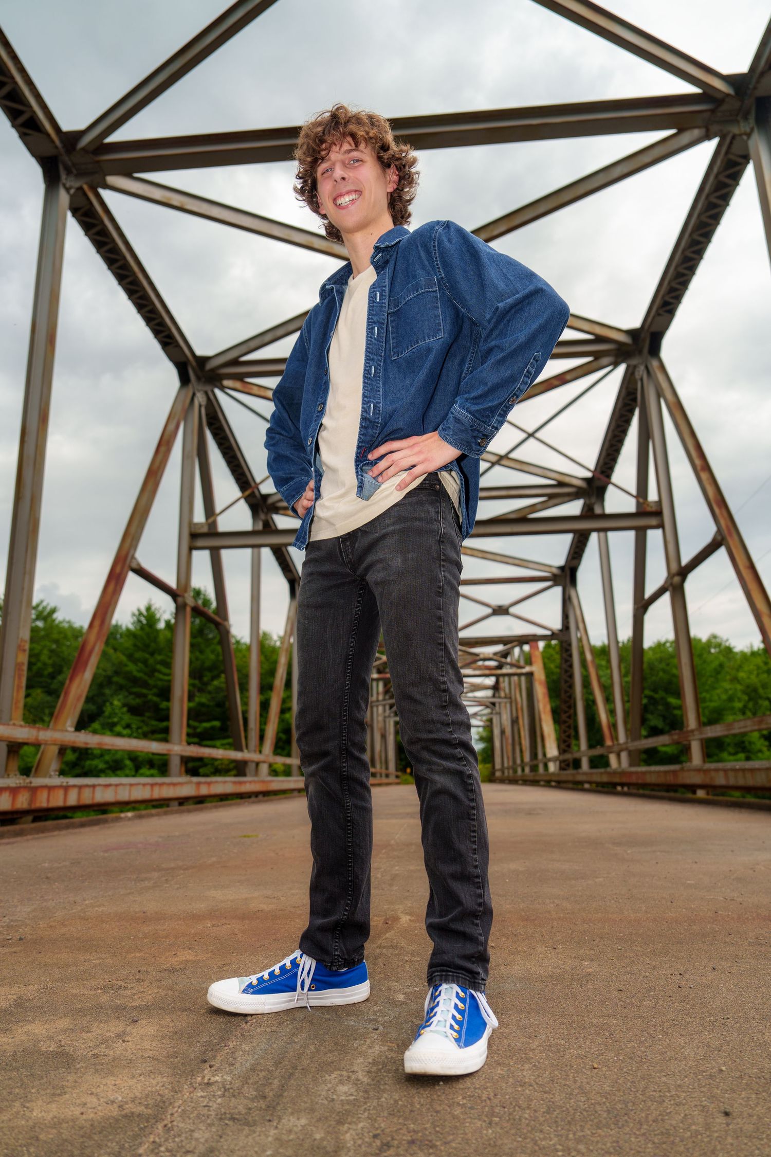 Young man with curly hair in denim shirt, black jeans, and blue Converse sneakers posing on a metal bridge.
