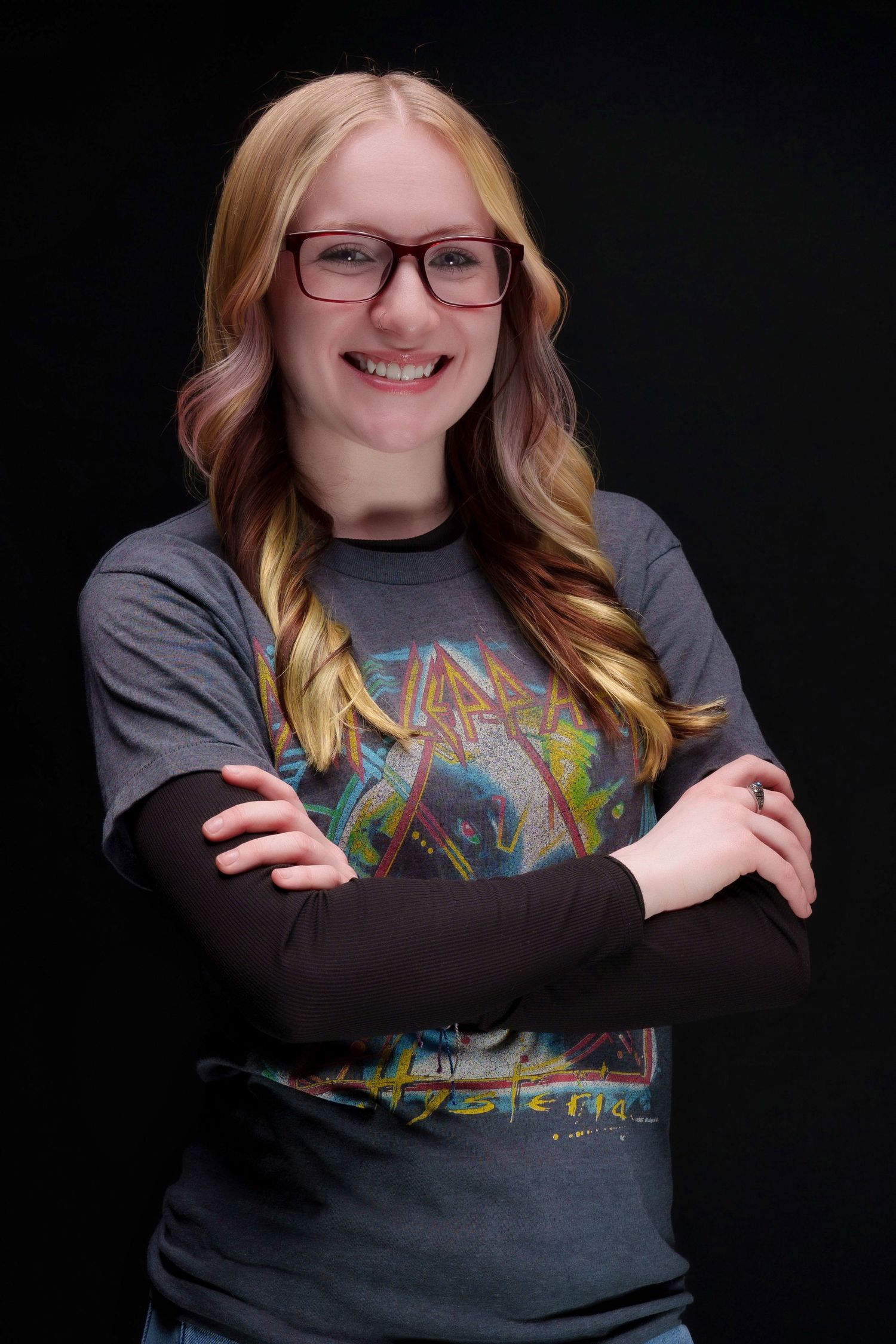 Smiling young woman with glasses and blonde hair wearing a Def Leppard t-shirt, arms crossed, against black background.