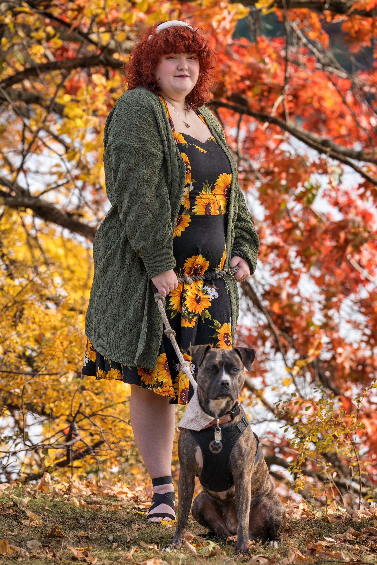 A person wearing a green jacket and sunflower garland stands with their dog among vibrant autumn foliage.