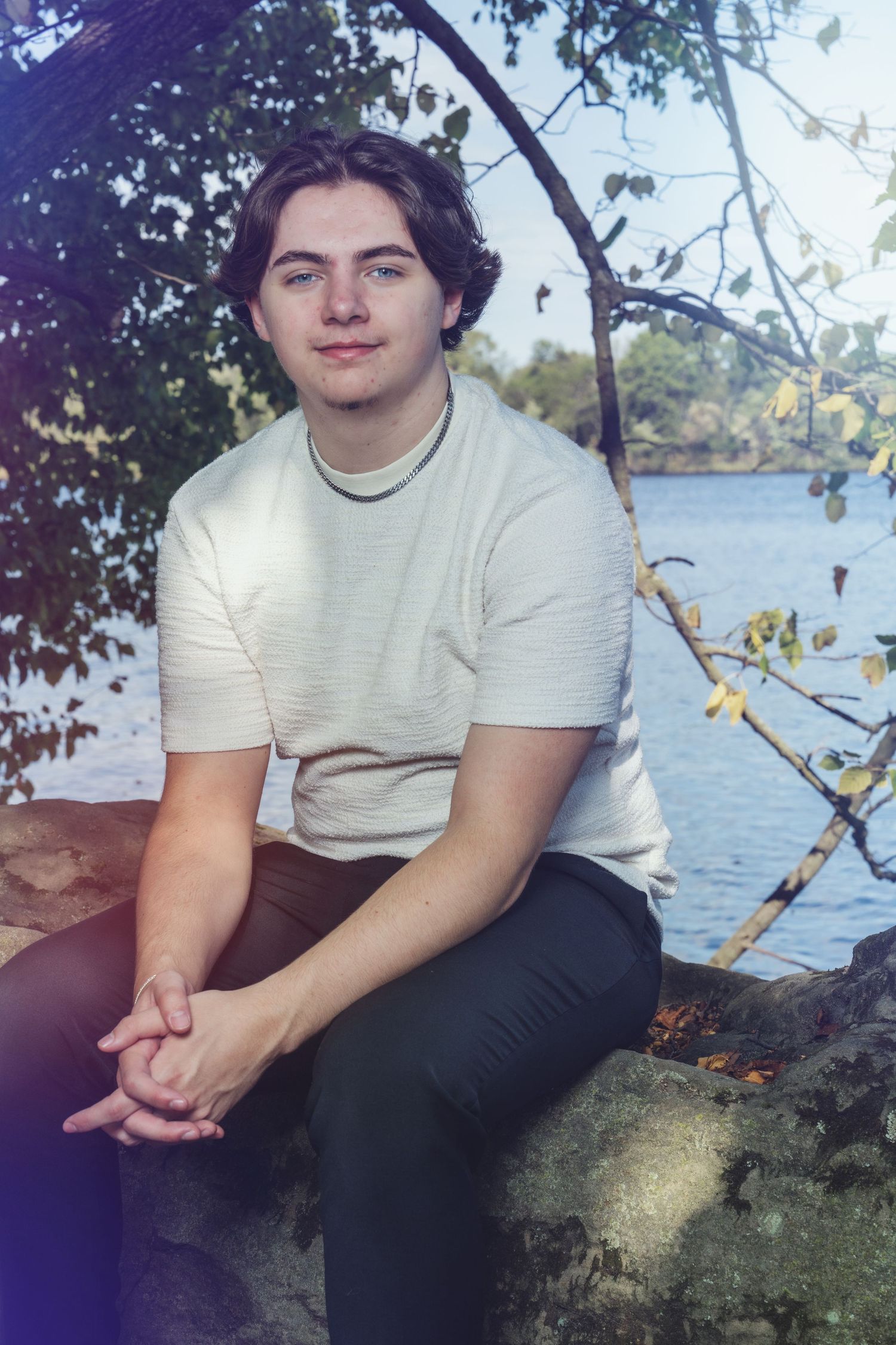 Person in a white t-shirt and dark pants sits by a waterfront with trees and foliage in the background.
