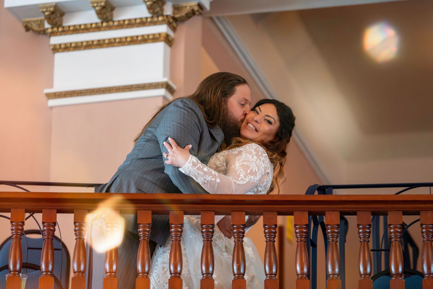 A couple shares an intimate moment while leaning over an ornate wooden railing in an elegant indoor setting.