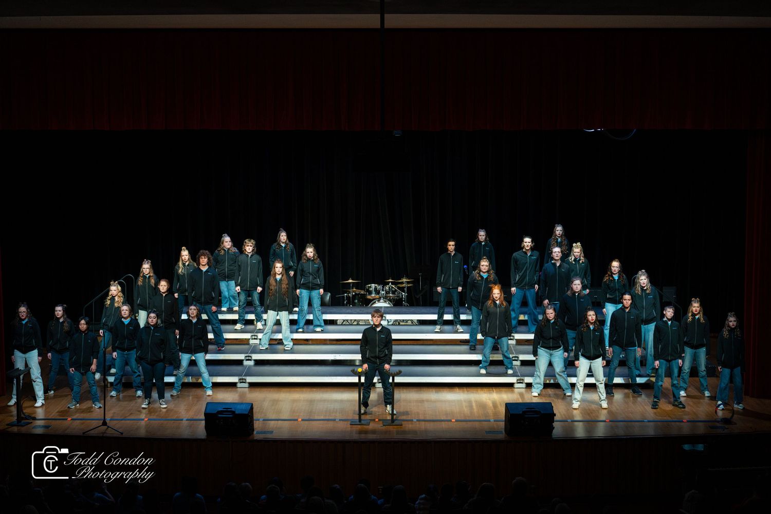 Performers in dark attire stand on illuminated risers during a stage production against a black background.