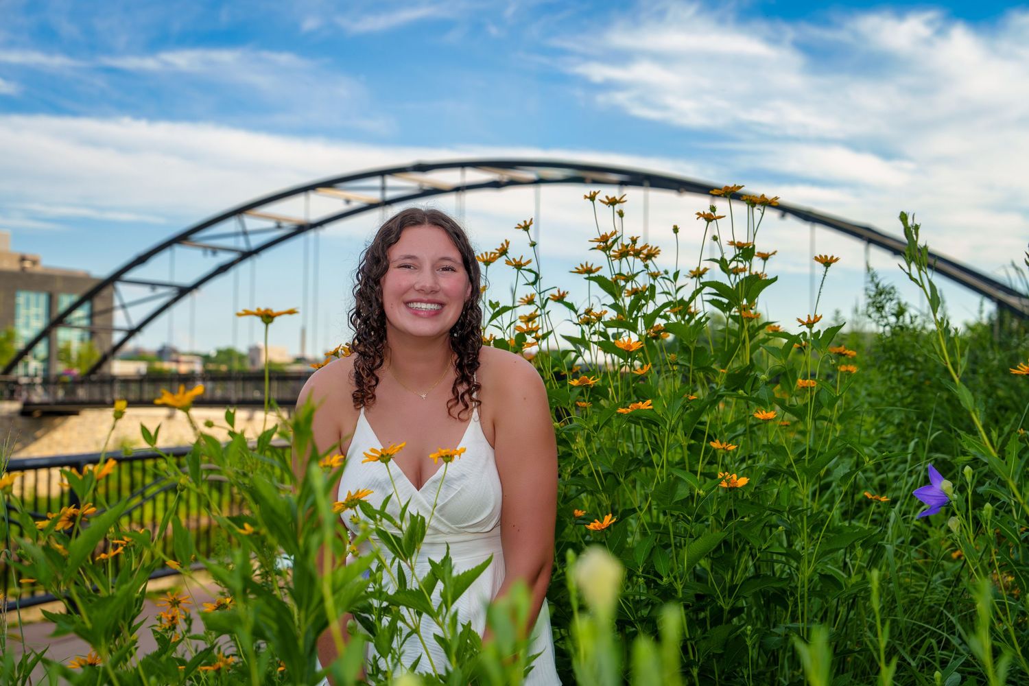 Smiling bride in white dress surrounded by wildflowers with an arched bridge and blue sky in the background.