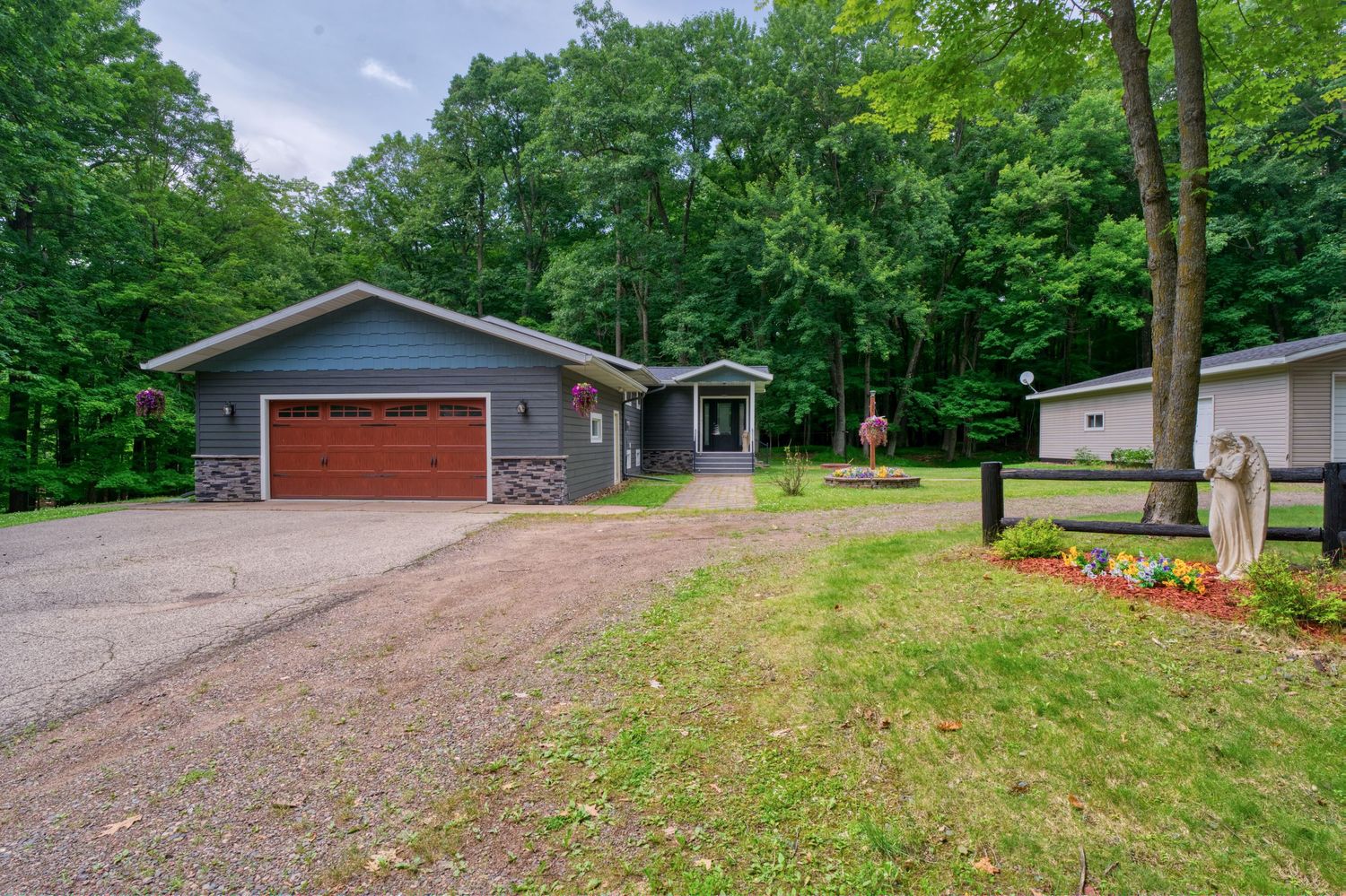 Gray ranch home with attached garage surrounded by lush green trees and manicured lawn in a wooded setting.
