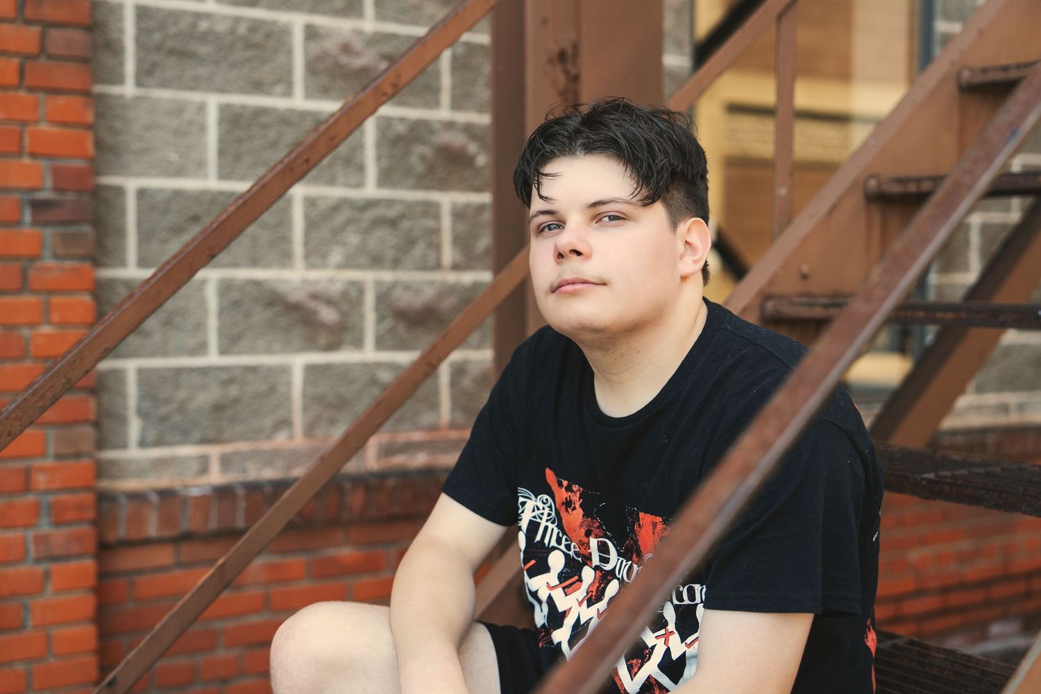 Person in black t-shirt with white design stands against brick wall with metal stairs in background.