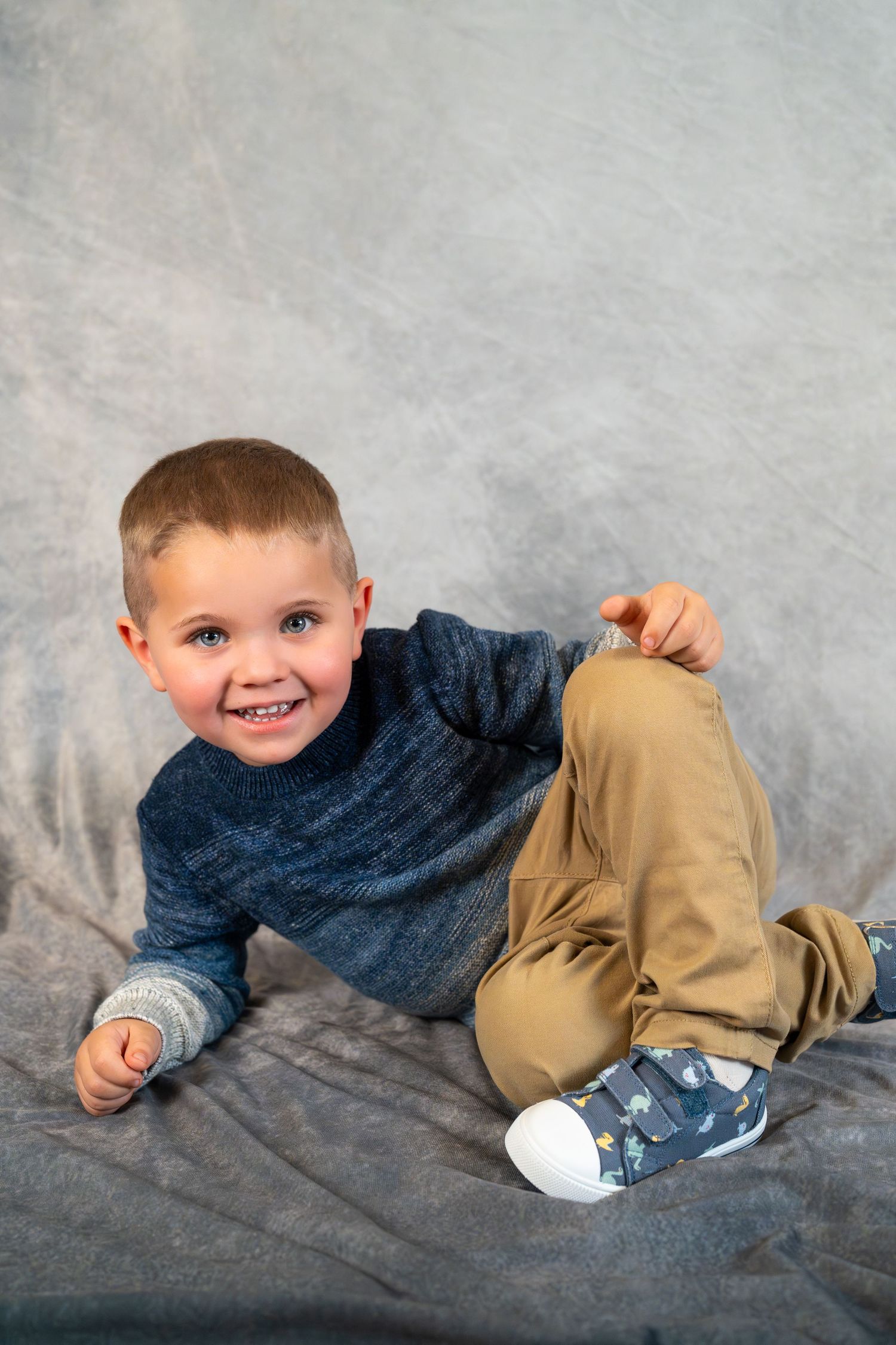 A young child in a blue sweater and khaki pants lies playfully on a gray backdrop while smiling.