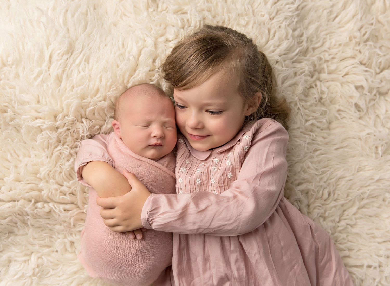 Two children in soft pink outfits cuddle together on a cream-colored fluffy blanket in a tender portrait. Toowoomba Photographer