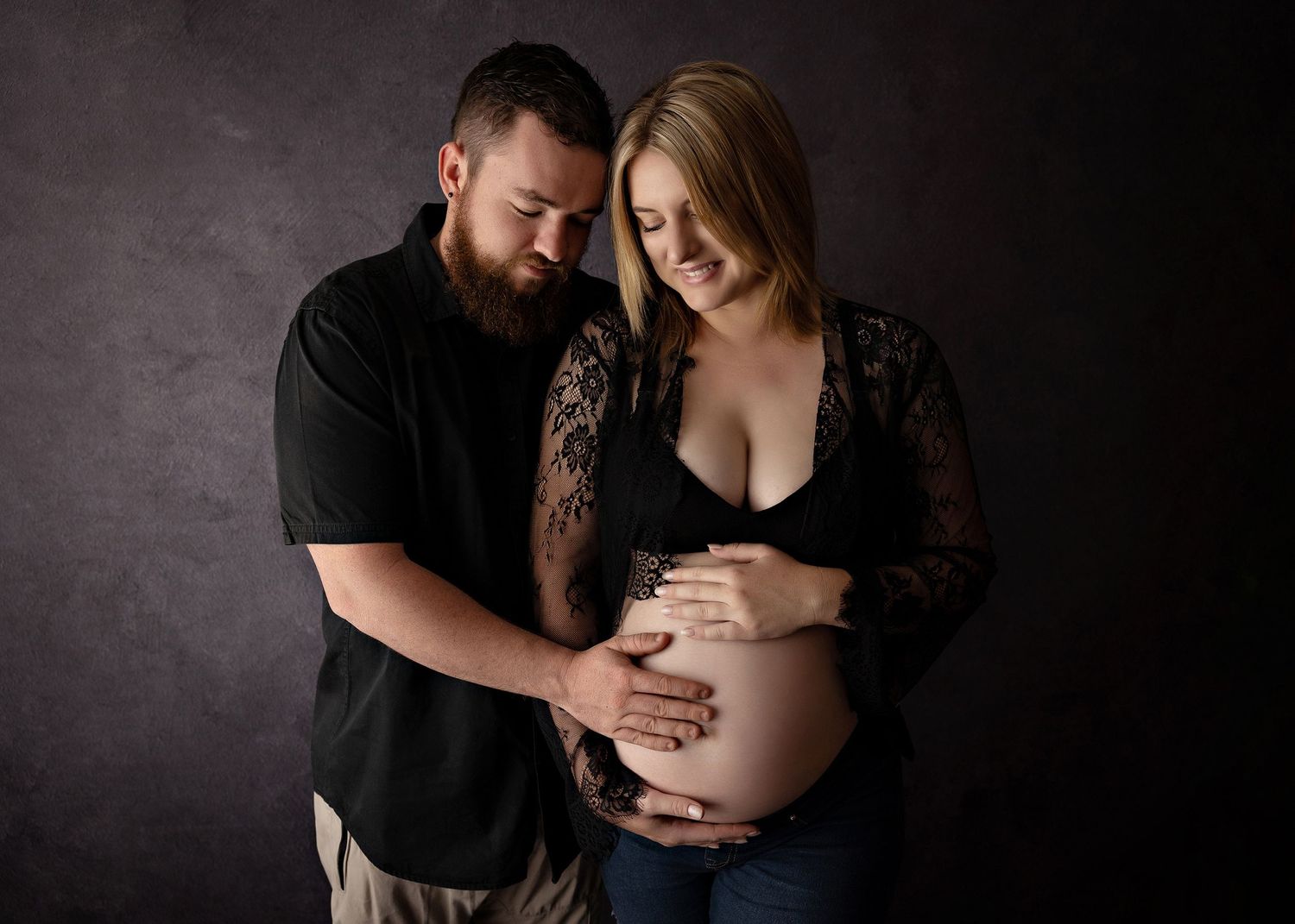 An intimate maternity photography session with a couple against a dark dramatic background.