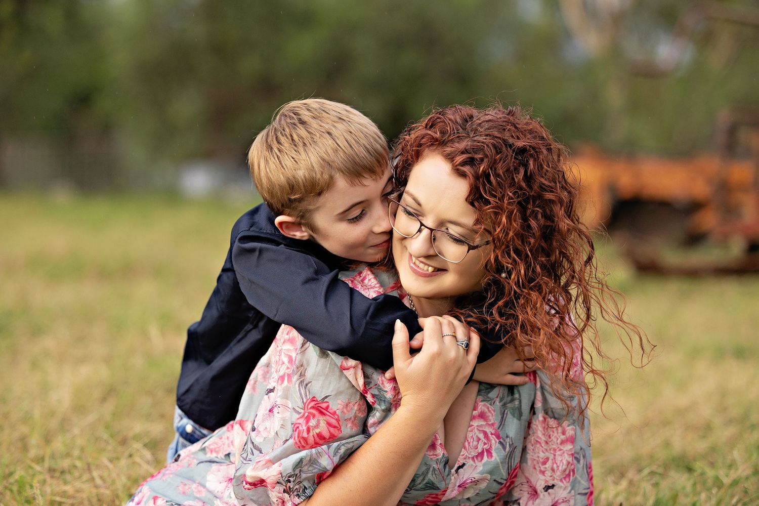 A young boy hugging a curly red-haired woman wearing glasses outdoors on green grass.