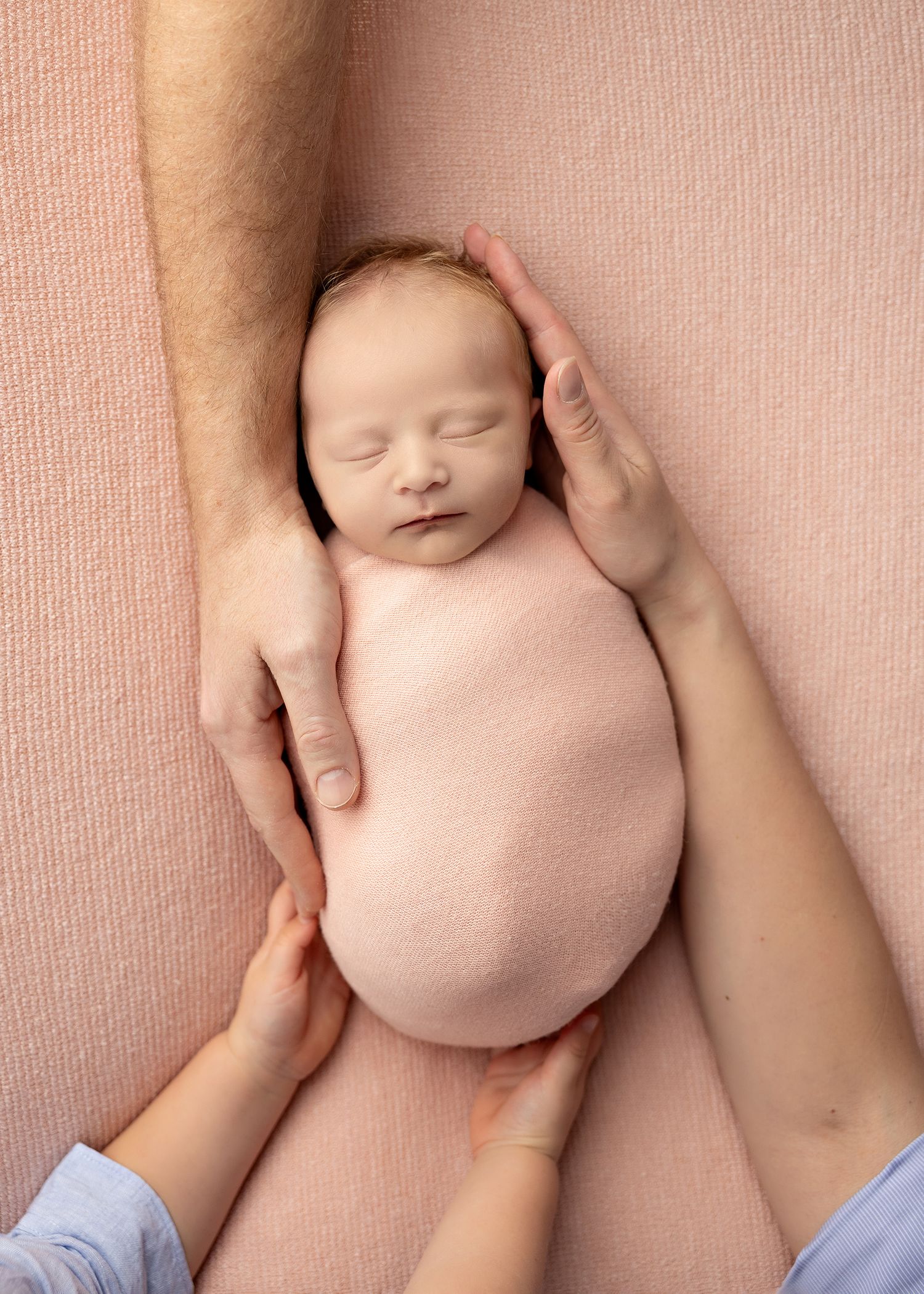 A series of hands gently massaging a sleeping newborn baby on a soft pink blanket.