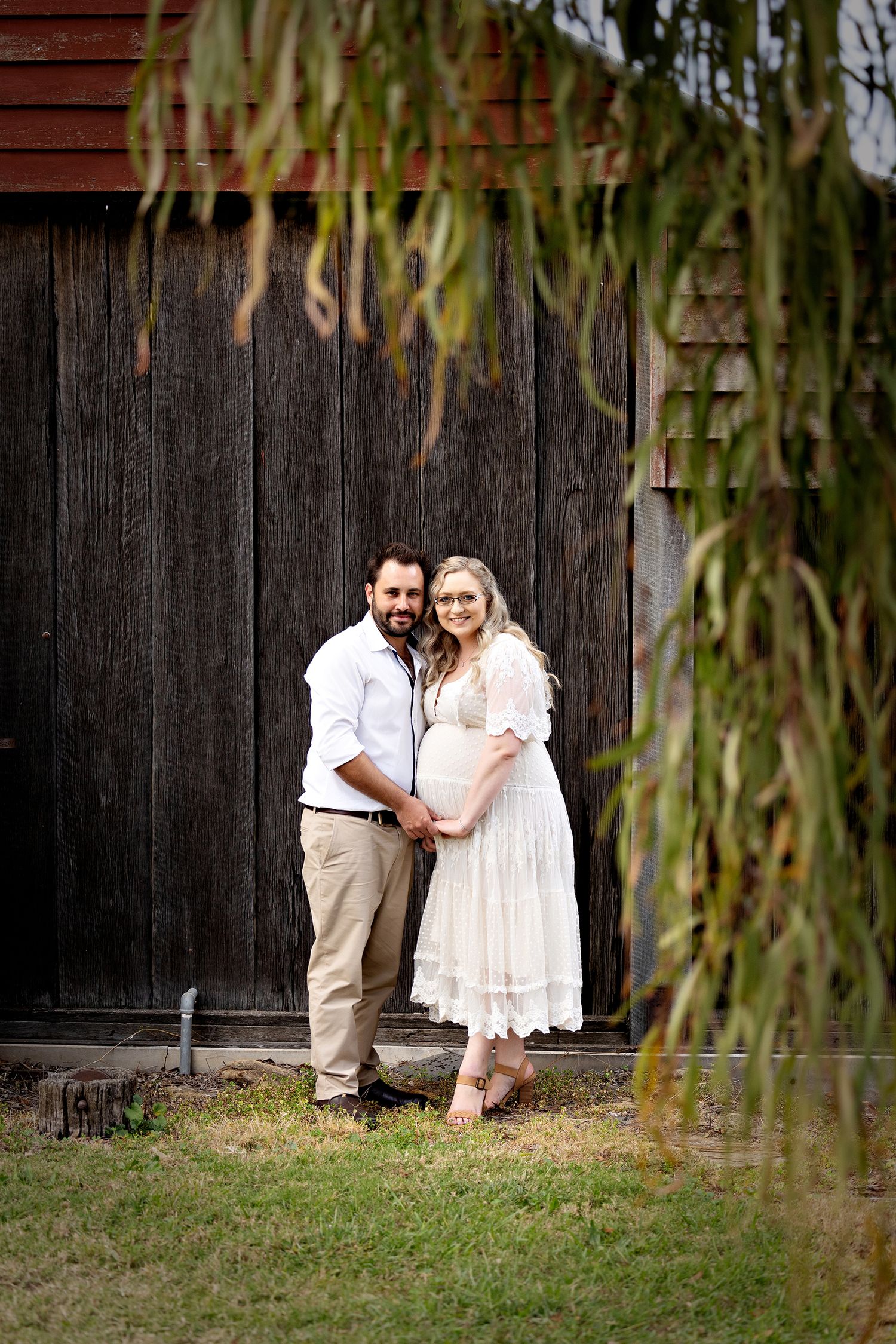 Expectant couple poses together against rustic barn wall with hanging willow branches.