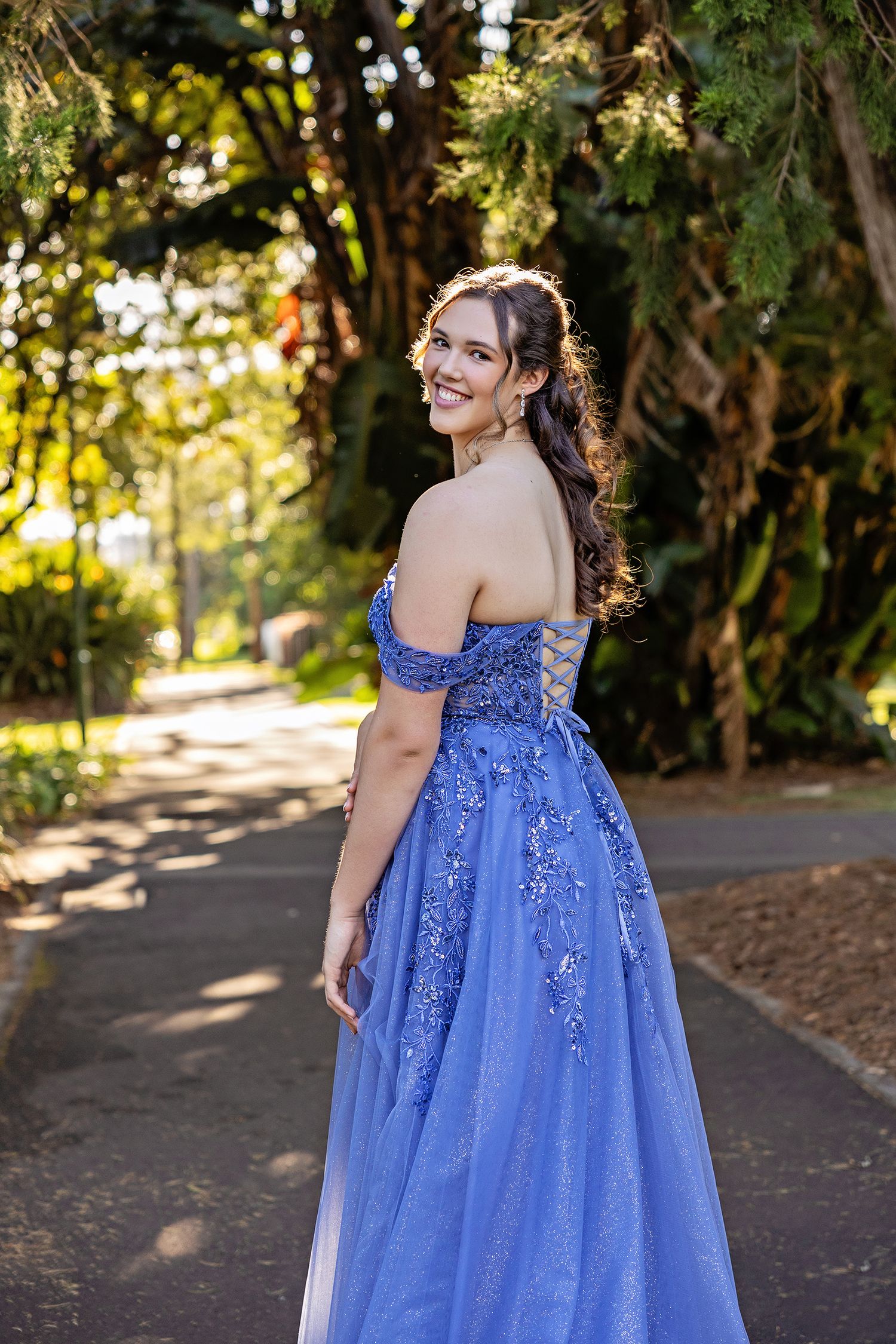 Young woman in blue off-shoulder floral sequin ball gown smiling outdoors on a path surrounded by lush greenery.
