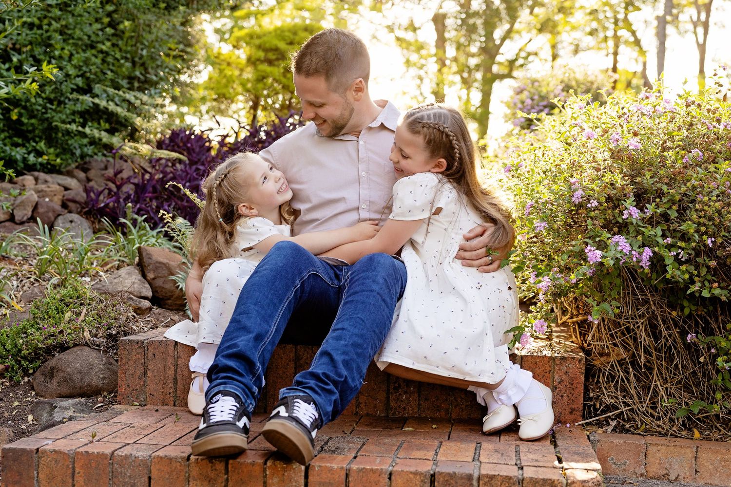 A father sits on brick steps with his two young daughters in white dresses, sharing a warm embrace in a garden.