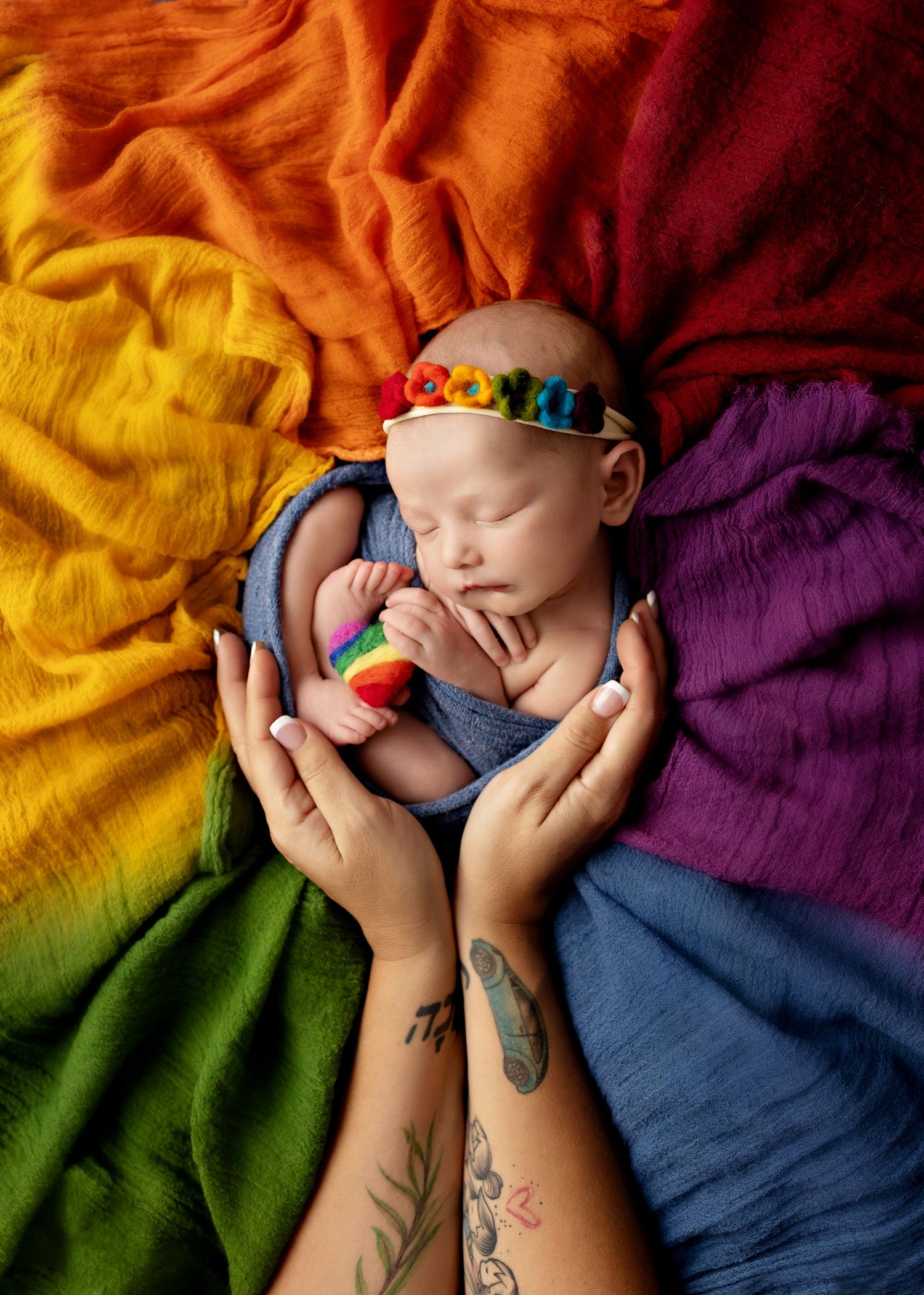 A sleeping infant curled up on colorful rainbow tie-dye fabric with a toy and visible tattoos in the frame.Toowoomba Photographer