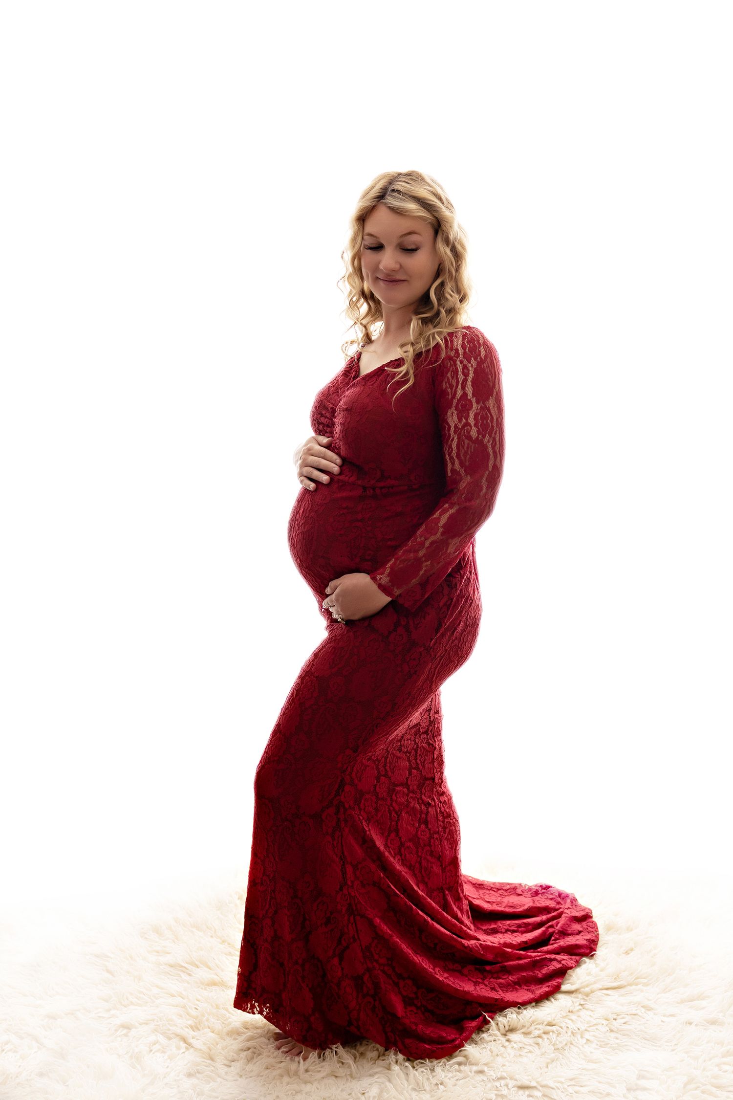 A pregnant woman in a flowing red lace dress poses gracefully on a white backdrop.