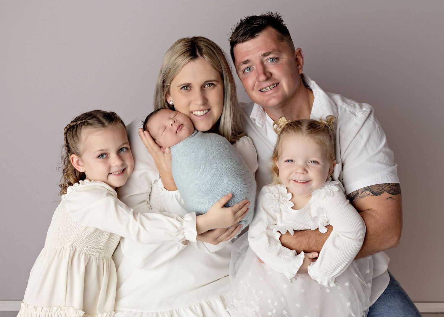 Family of five posing for a professional portrait, parents holding newborn baby and toddler, with older daughter smiling.
