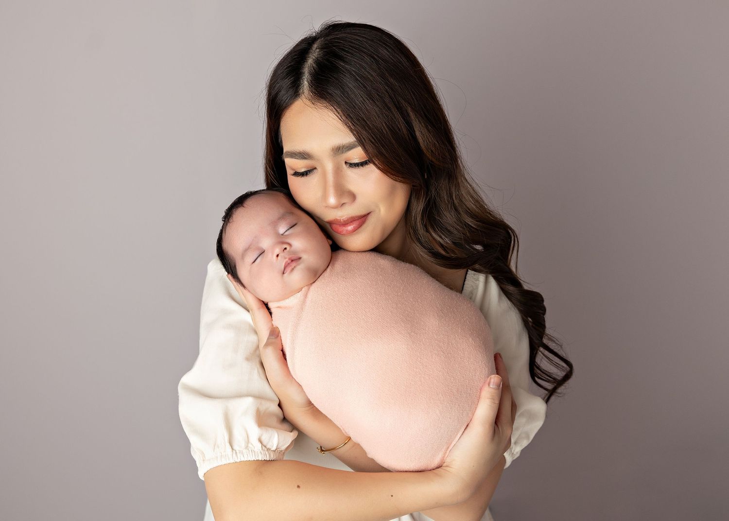 A tender newborn portrait shows a sleeping baby being gently cradled against a soft cream backdrop.