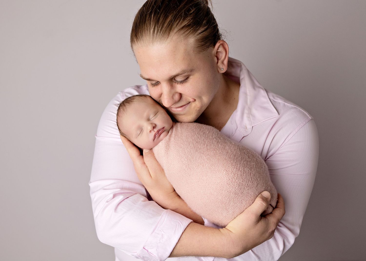 A father tenderly holds his sleeping newborn baby wrapped in a soft pink blanket during a studio portrait session.