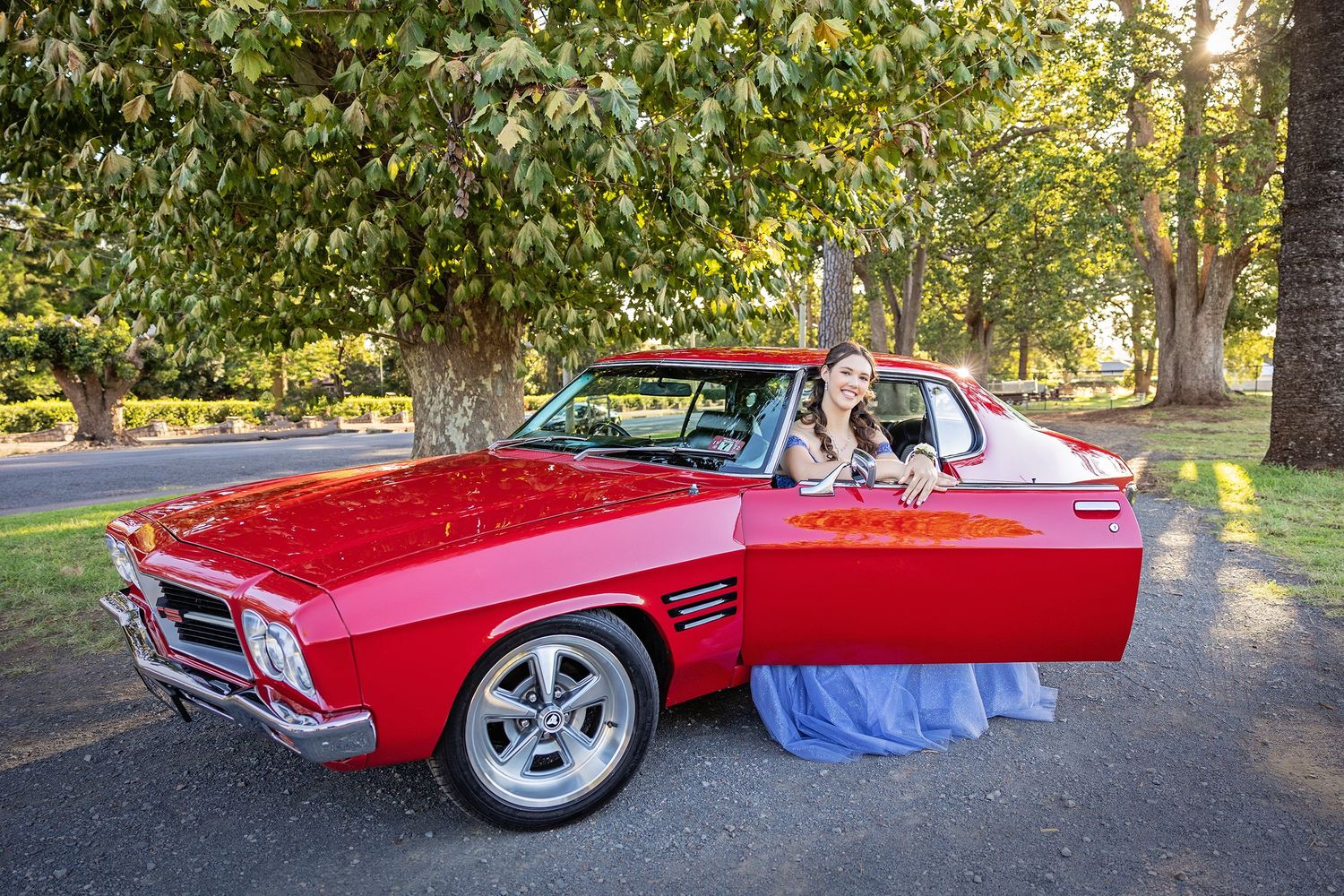 Bride in blue gown posing beside a classic red Holden muscle car under lush green trees on a scenic driveway.