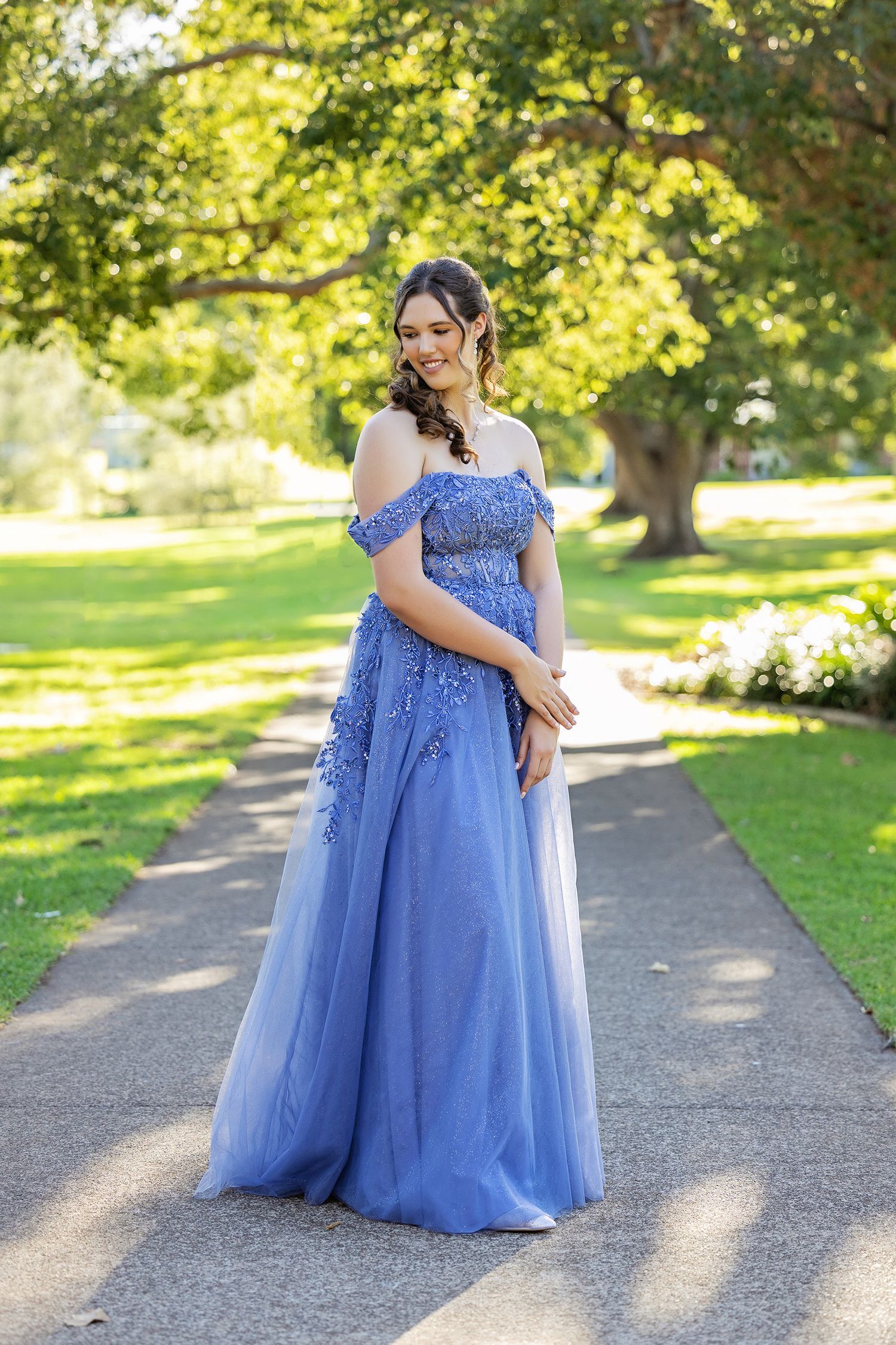 Young woman in a blue off-shoulder floral ball gown poses on a garden path surrounded by lush green trees.