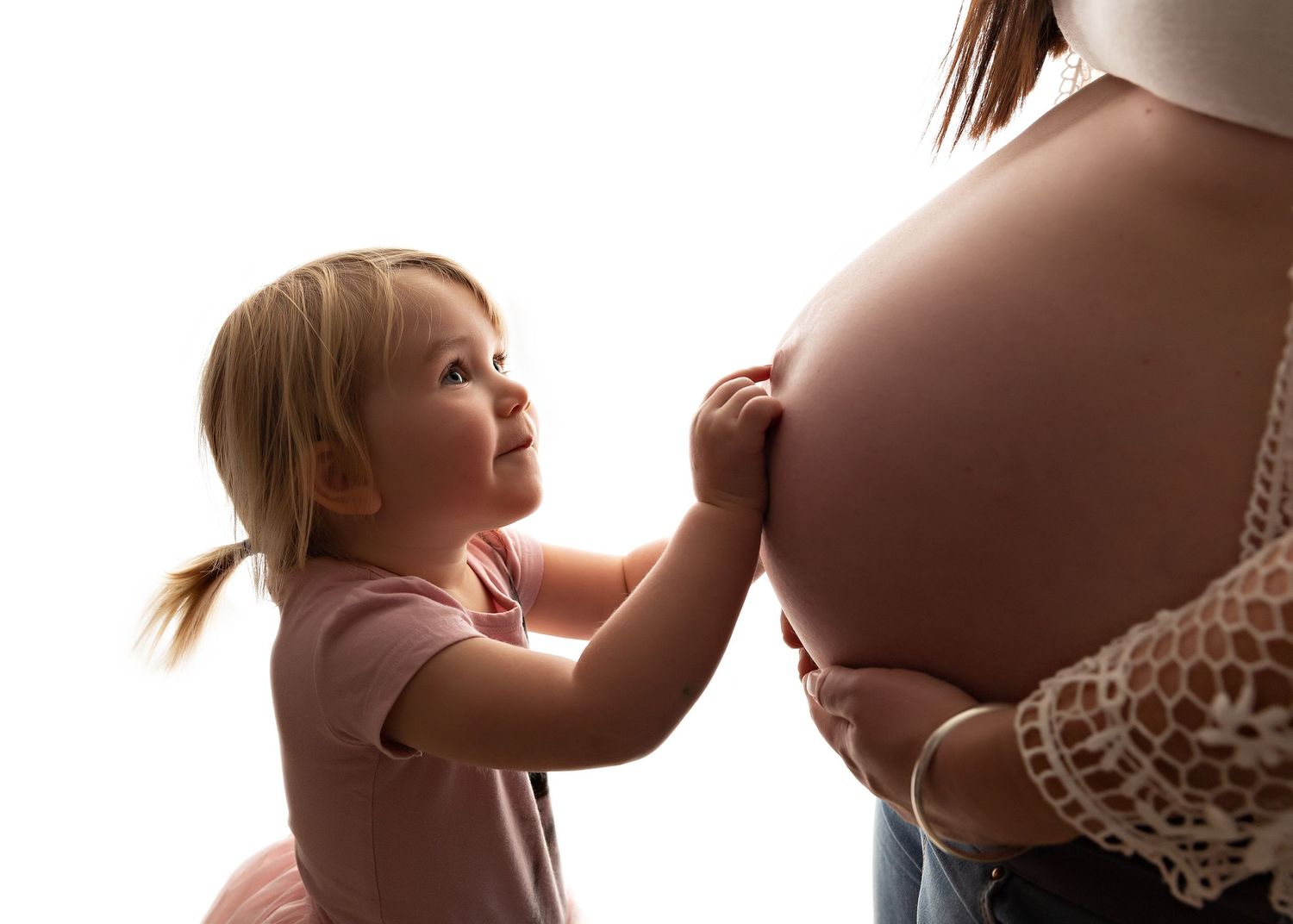 Young child touching pregnant belly in tender moment against backlit background.
