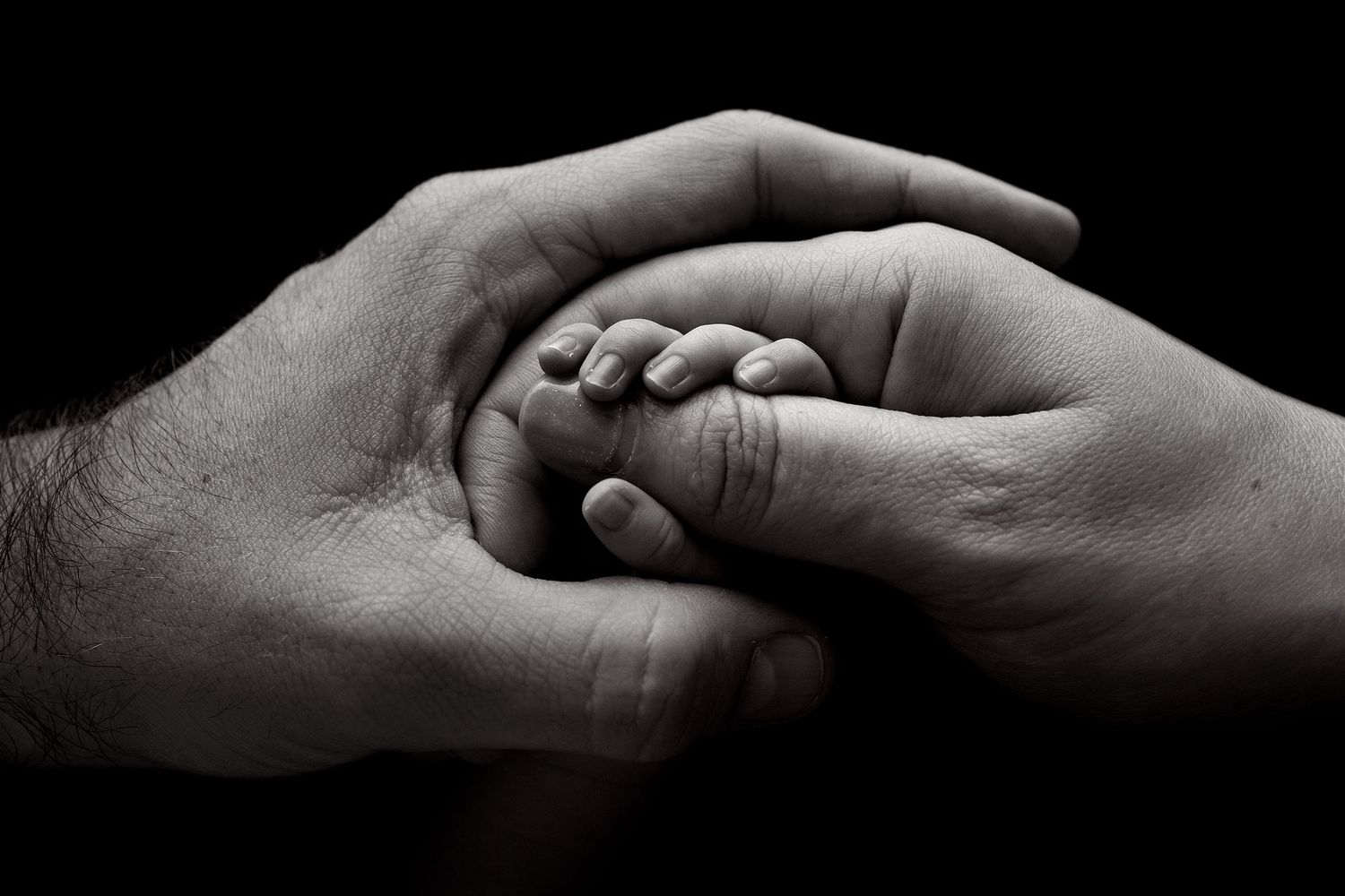 Black and white photo of a baby's tiny hand gently held between two adult hands, symbolizing love and protection.